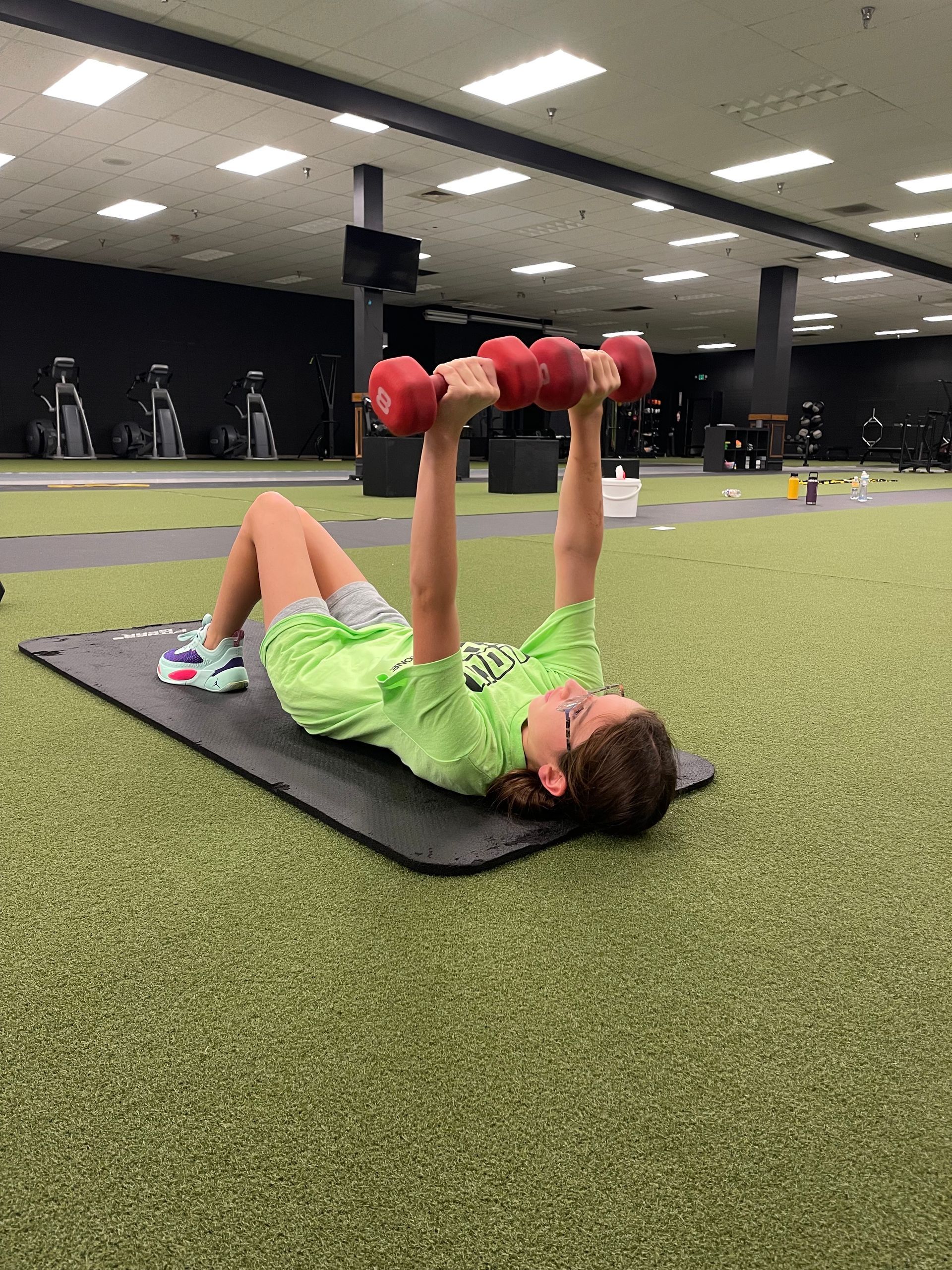 A woman is laying on a mat in a gym lifting dumbbells.