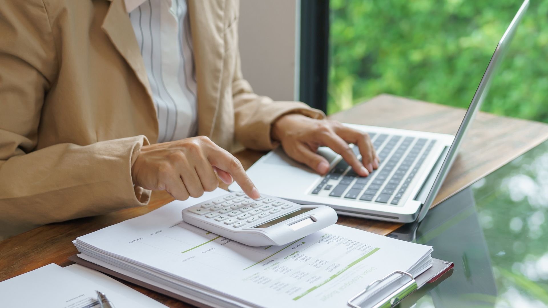 Person in tan blazer using calculator and laptop, paperwork on desk.