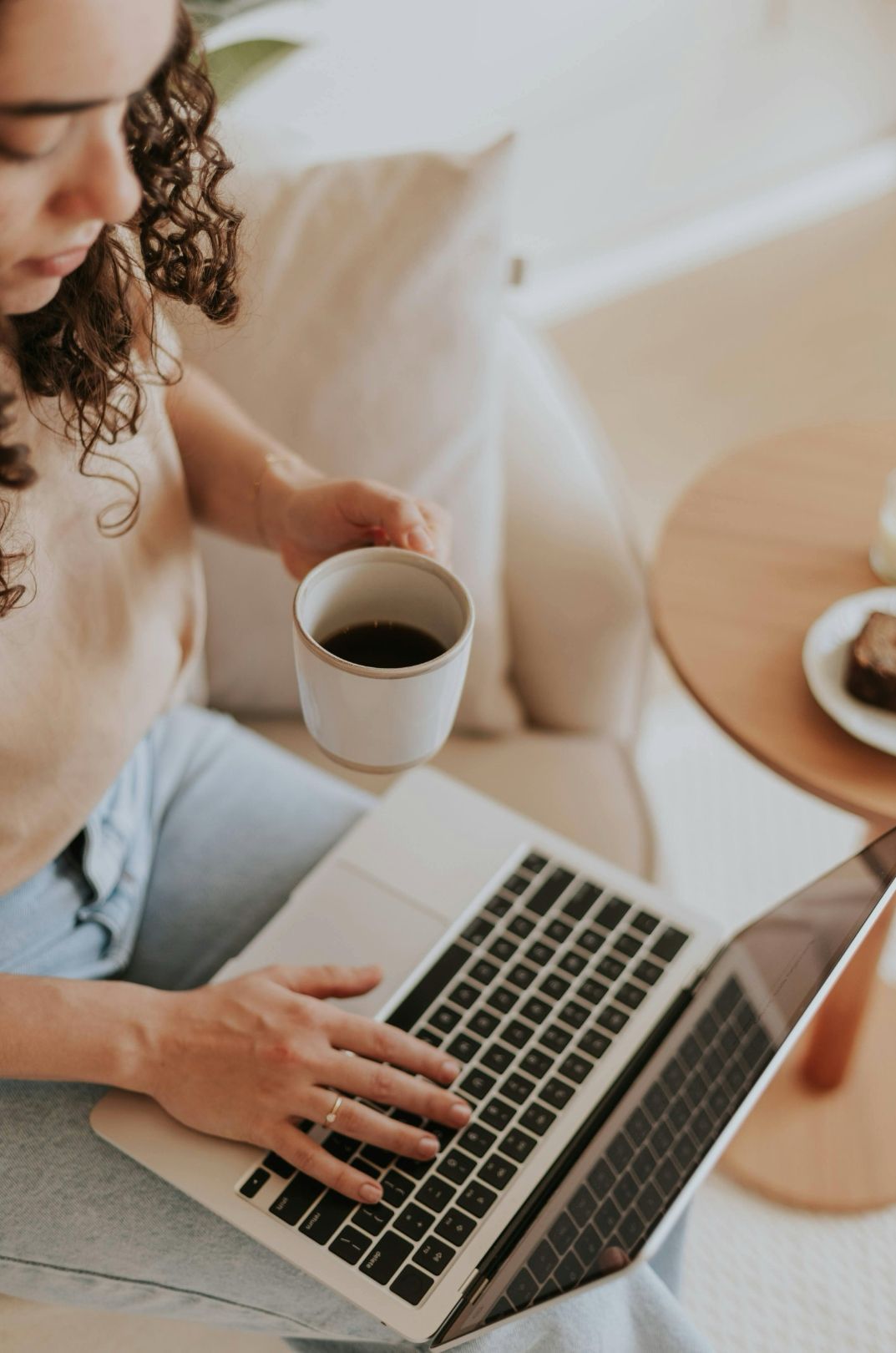 Woman sitting on a couch, using a laptop, holding a mug of coffee.