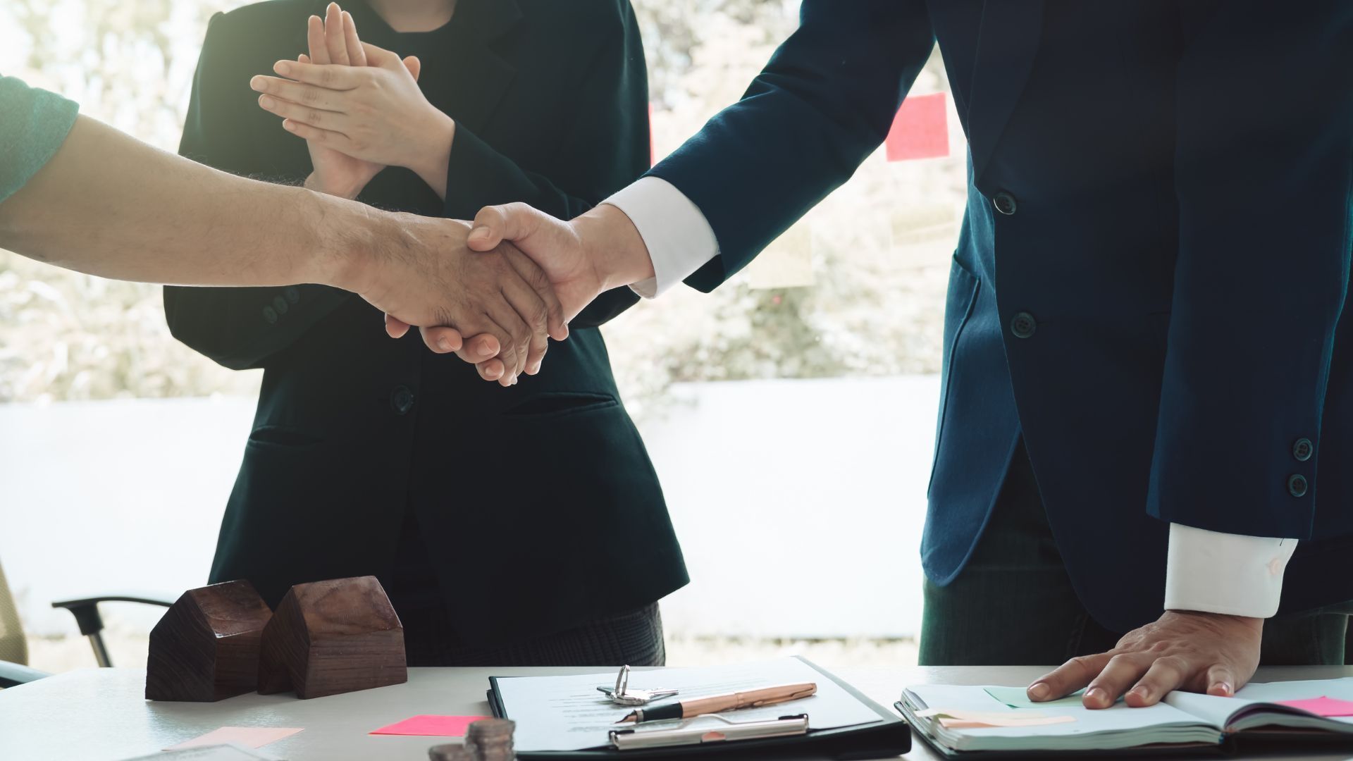 Two business professionals shaking hands over a desk with model houses and documents, while a third person applauds.