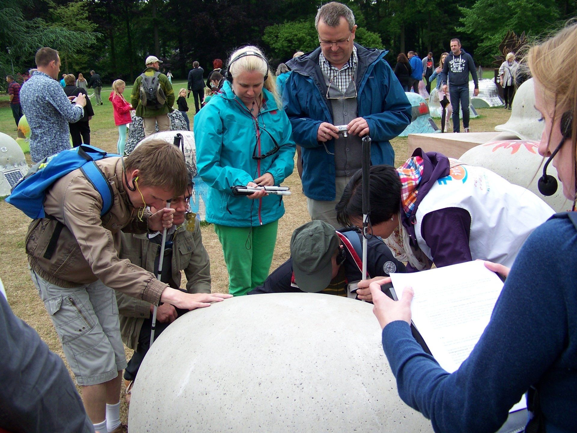 Enkele blinde en slechtziende bezoekers hebben zich rond een betonnen helm verzameld. Ze tasten de helm af terwijl Susanne uitleg geeft.
