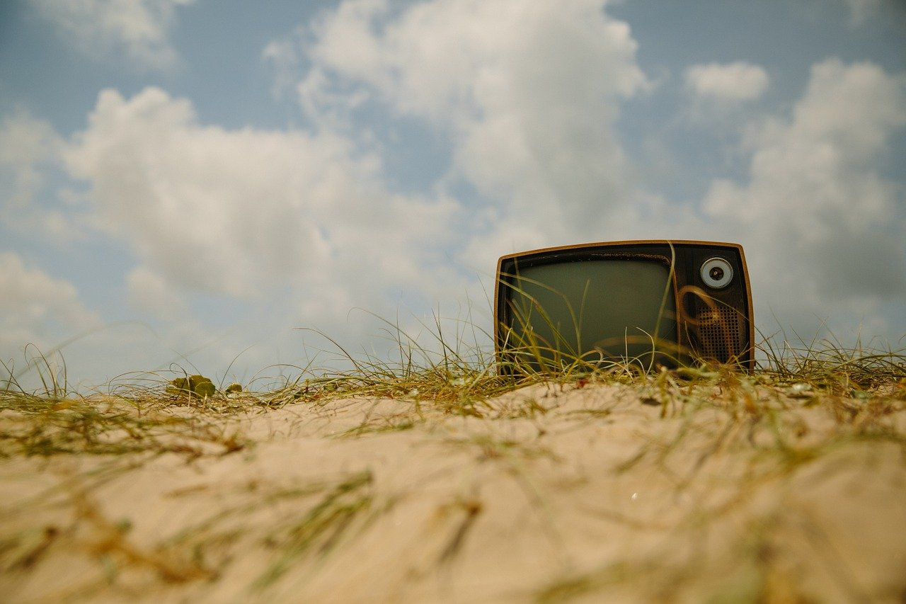 Een ouderwetse tv ligt in een duinlandschap met op de achtergrond enkele wolkenplukjes