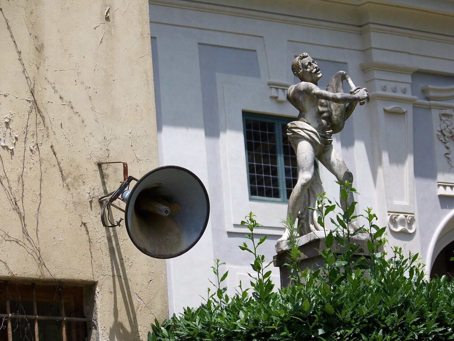 Een Italiaanse tuin met een oud standbeeld op een sokkel en daarnaast een luidspreker aan de muur van een gebouw…
