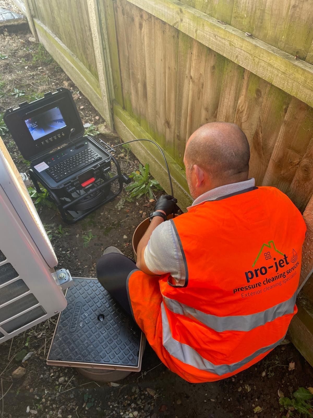 Man in orange vest inspects pipes with a camera, next to a fence and equipment.