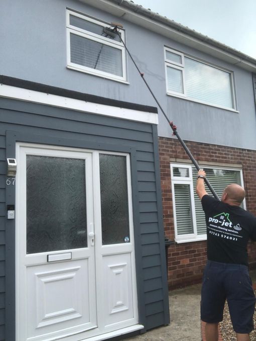 Man using a water-fed pole to clean a window on a two-story house with a blue exterior.