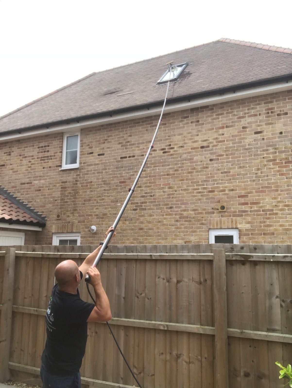 Man cleaning a window with a long-handled brush from behind a wooden fence. Building in the background.