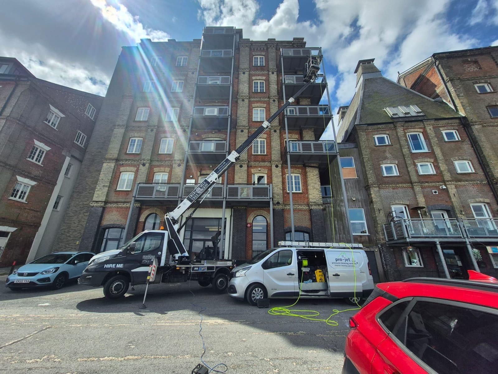 A crane truck is working on a brick building with balconies. Vehicles parked nearby.