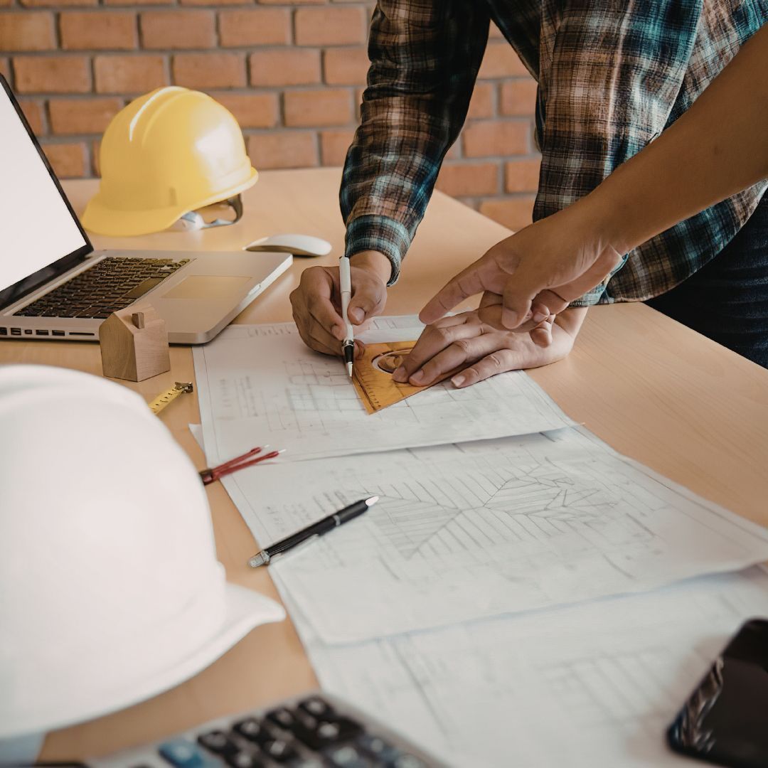 A construction manager wearing a white hard hat sitting at a wooden desk, carefully drawing on new home blueprints using a stencil and pencil.