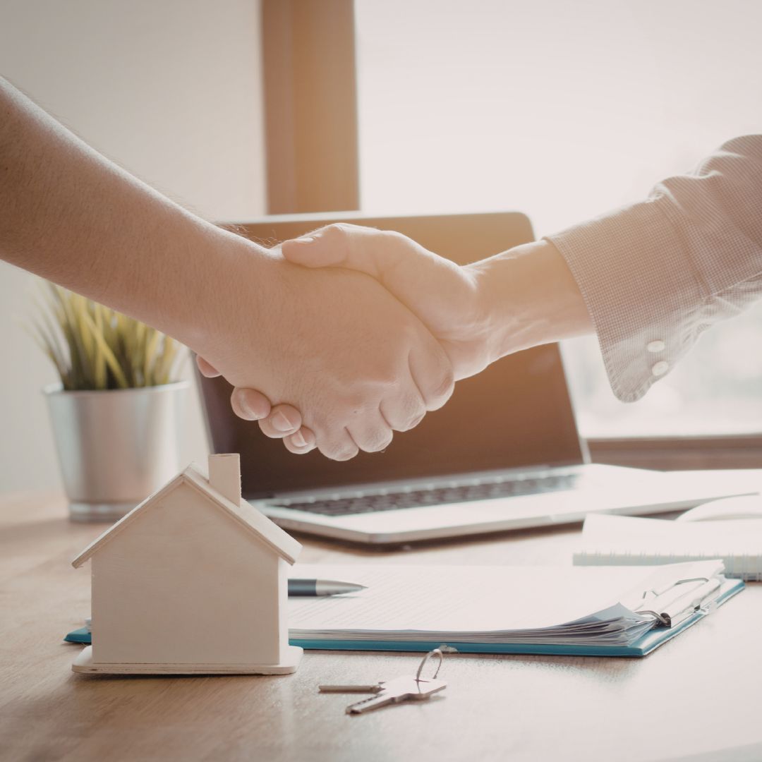 Close up of a real estate agent and a client shaking hands over a desk, with a set of house keys and a small model home resting on the table, representing a successful new construction closing.