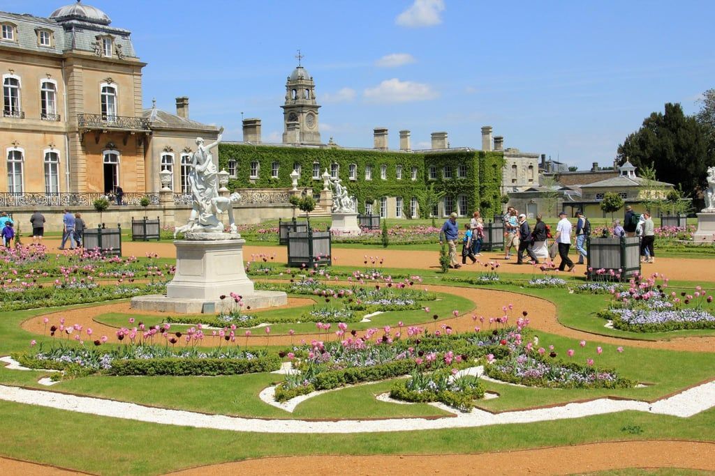 A group of people are walking in a garden with a large building in the background.