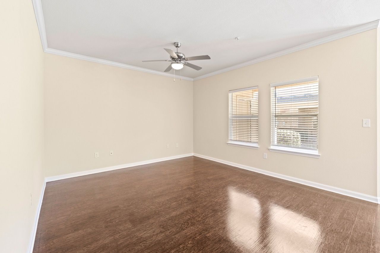 Living room in an apartment with beige walls, wood-like flooring, two windows with blinds, and a ceiling fan.