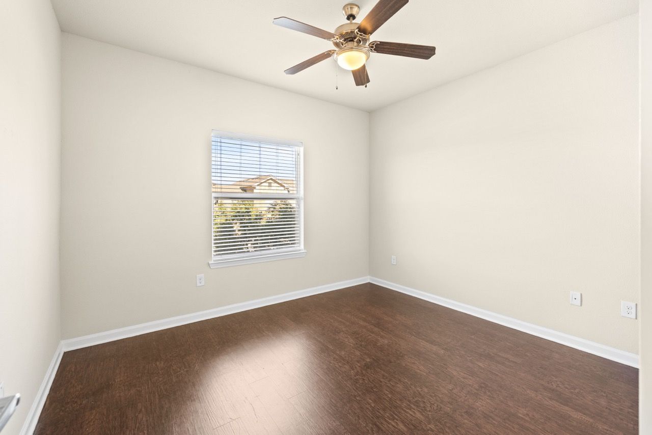 Empty bedroom with beige walls, dark wood-like flooring, a window with blinds, and a ceiling fan.