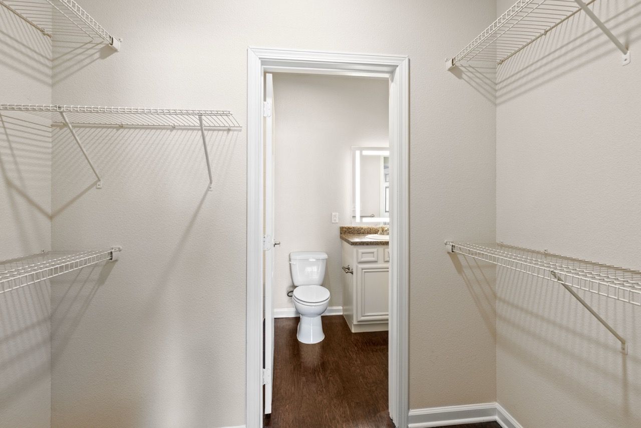 Walk-in closet with white wire shelves on both sides, opening to a bathroom with a toilet and vanity.