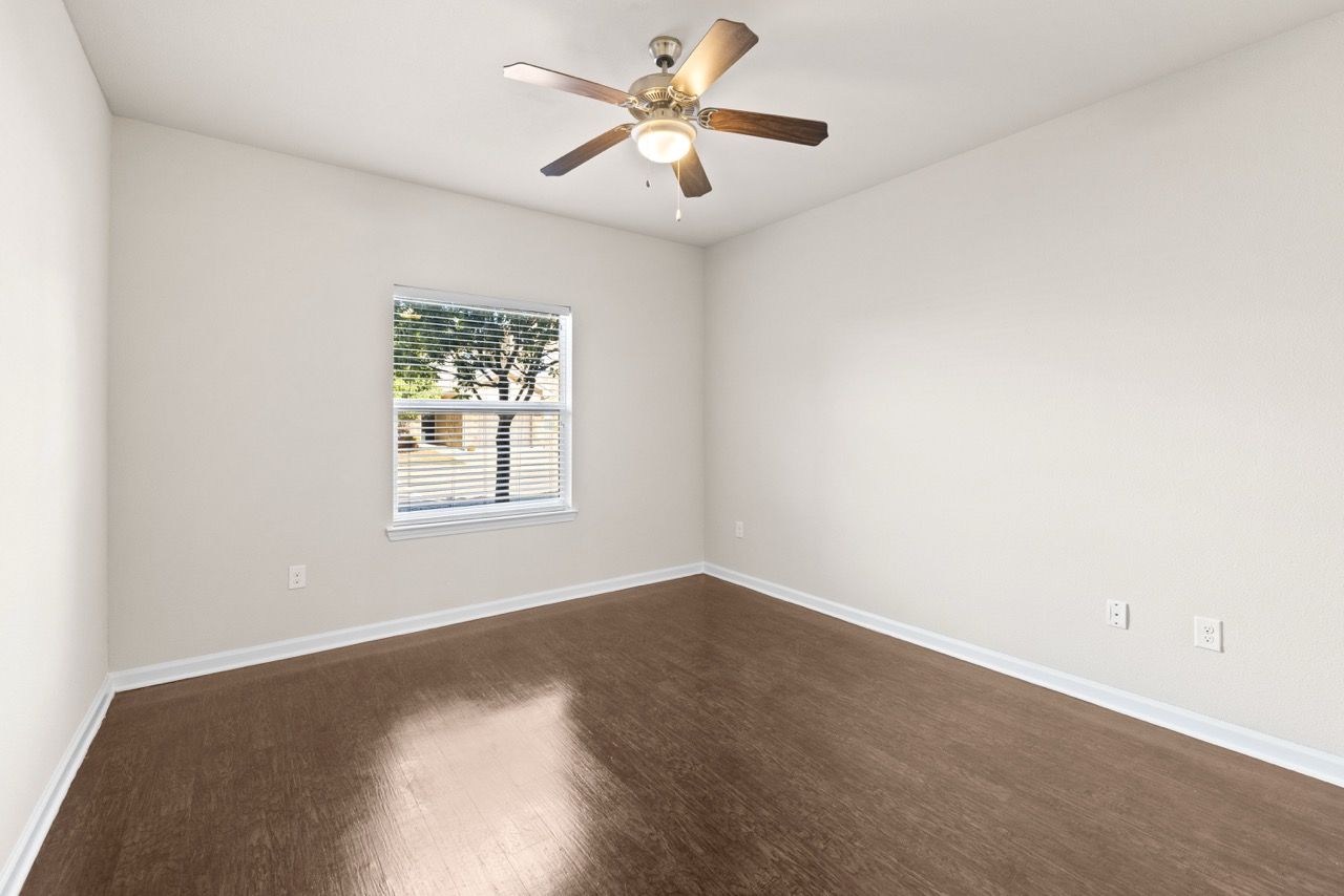 Empty beige bedroom with a window, ceiling fan, and brown wood-like flooring.