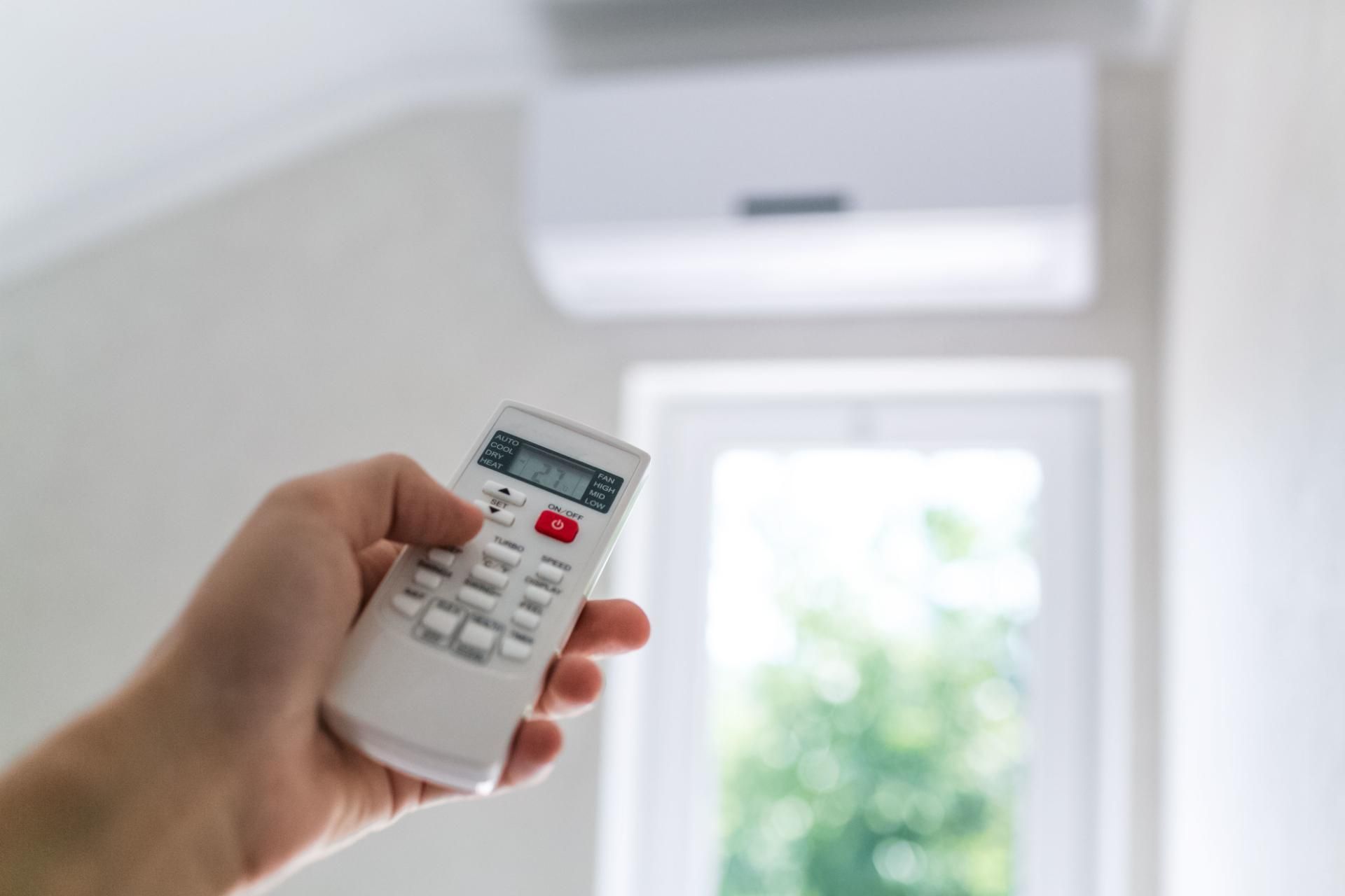 Mano che tiene il telecomando dell'aria condizionata, indicando l'unità montata a parete, pareti bianche, finestra soleggiata sullo sfondo.