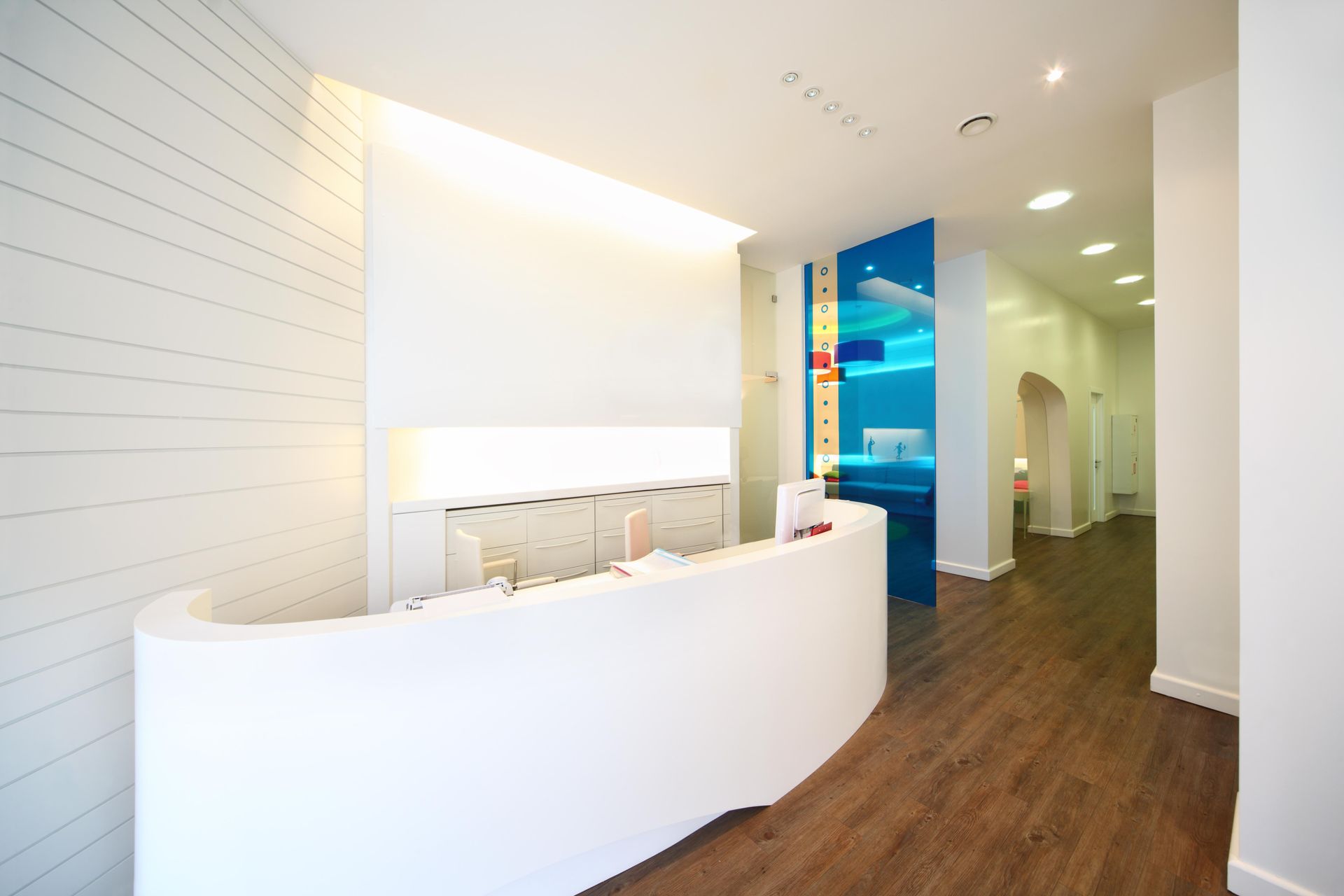 Modern white dental office reception area with curved desk, wood floors, and blue accent.