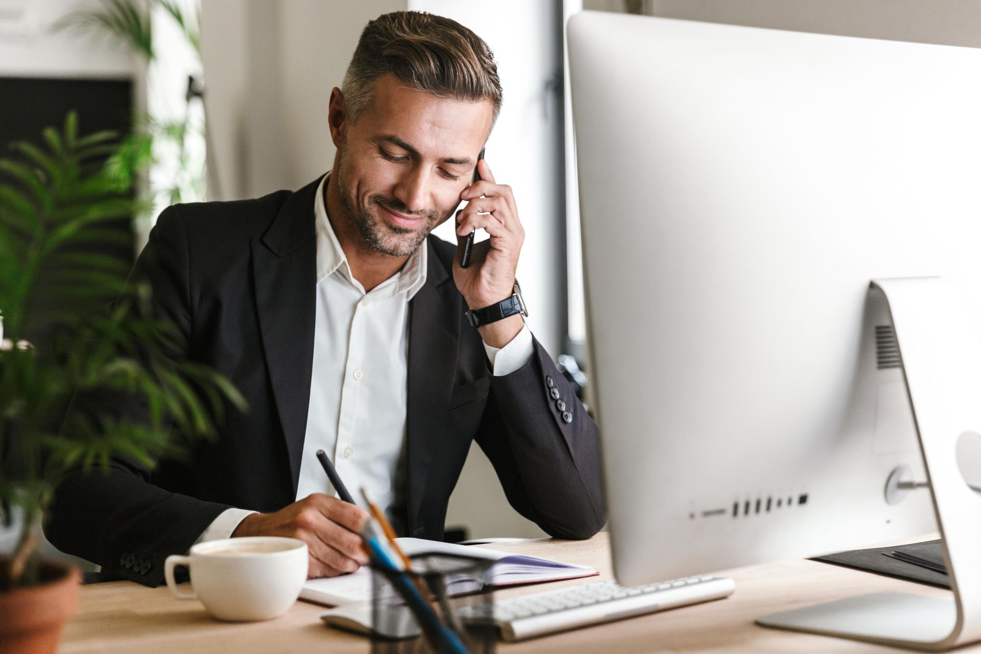 Man in suit on phone, writing at desk with computer and plant.