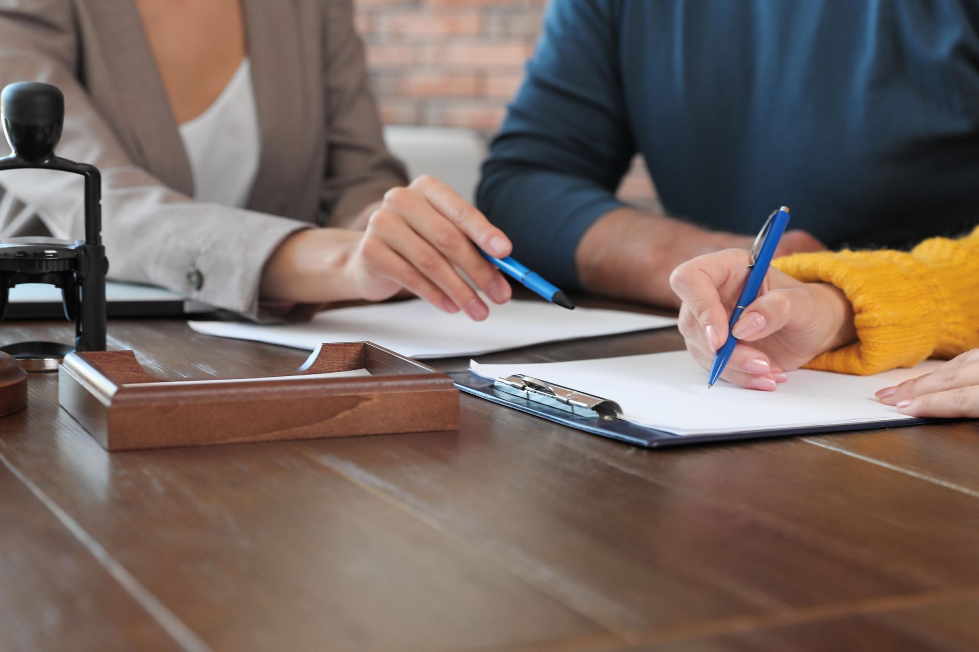People signing documents at a table. A person in a blazer holds a pen while another person signs. A stamp sits nearby.
