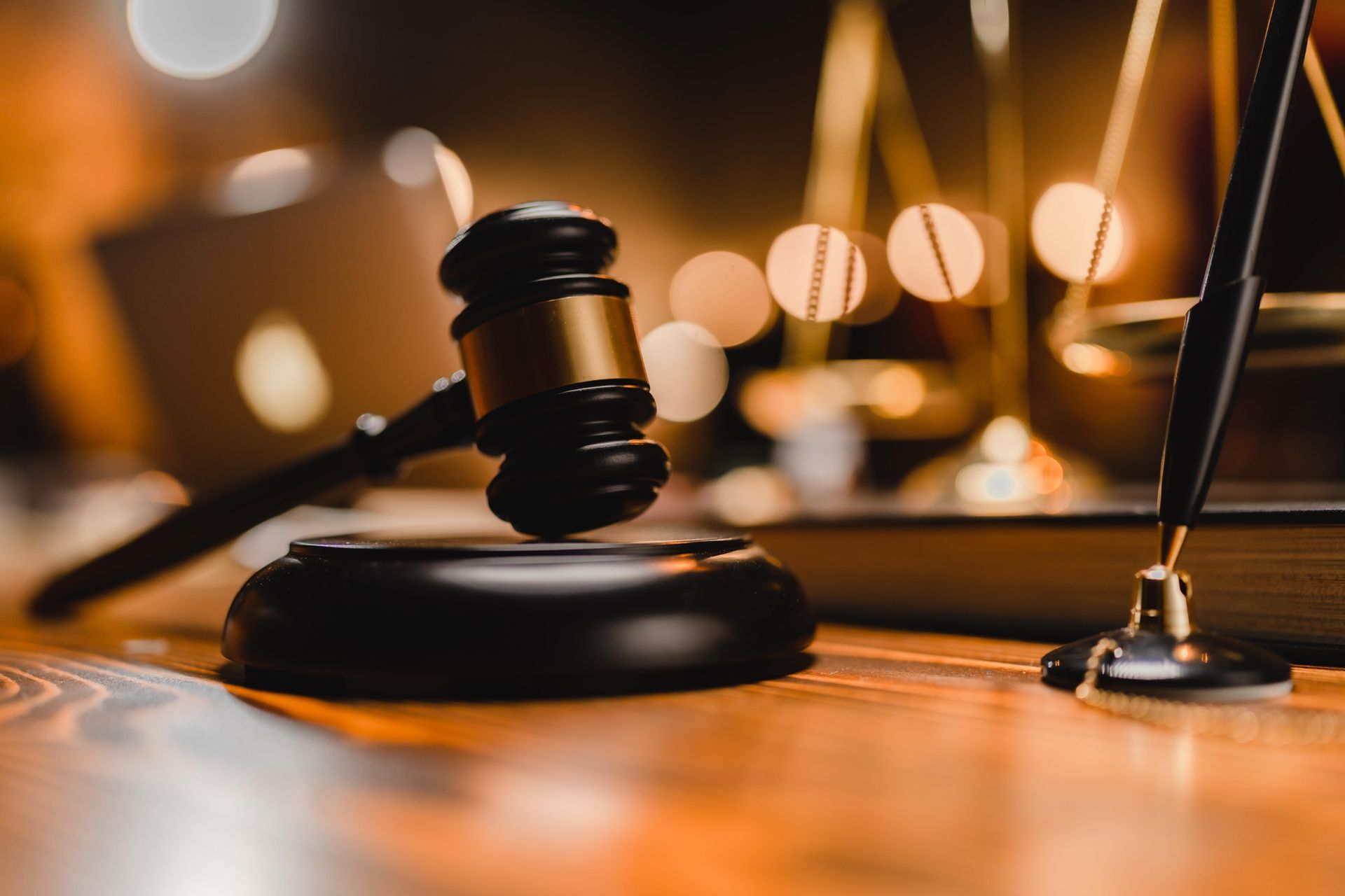 Gavel on a wooden desk with a balance scale and a book in the background, likely in a courtroom.