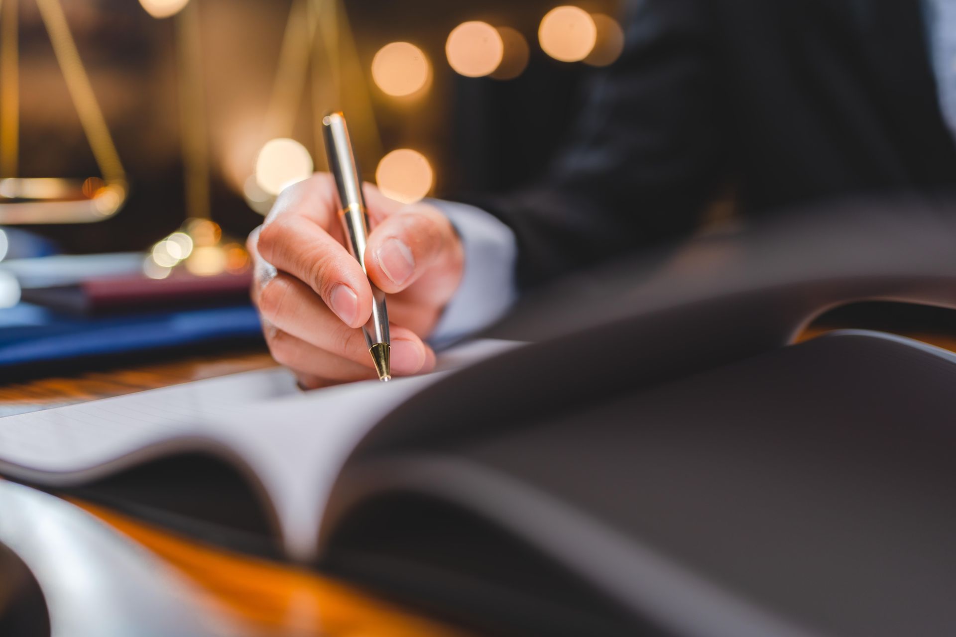 Person in suit writing with a pen in a book, scales of justice and blurred lights in background.