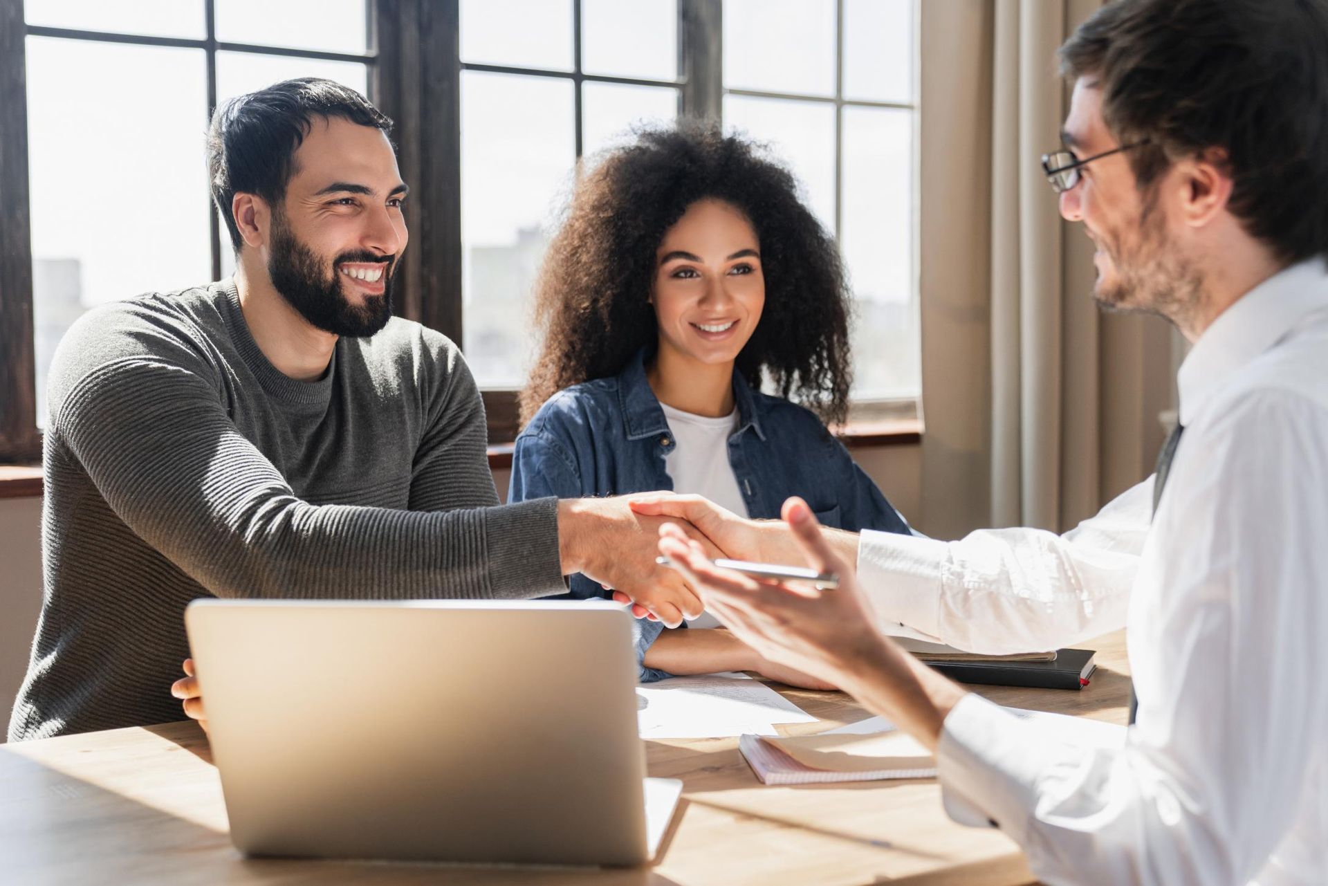 Couple shaking hands with a person at a table, likely after a deal, with a laptop and documents.