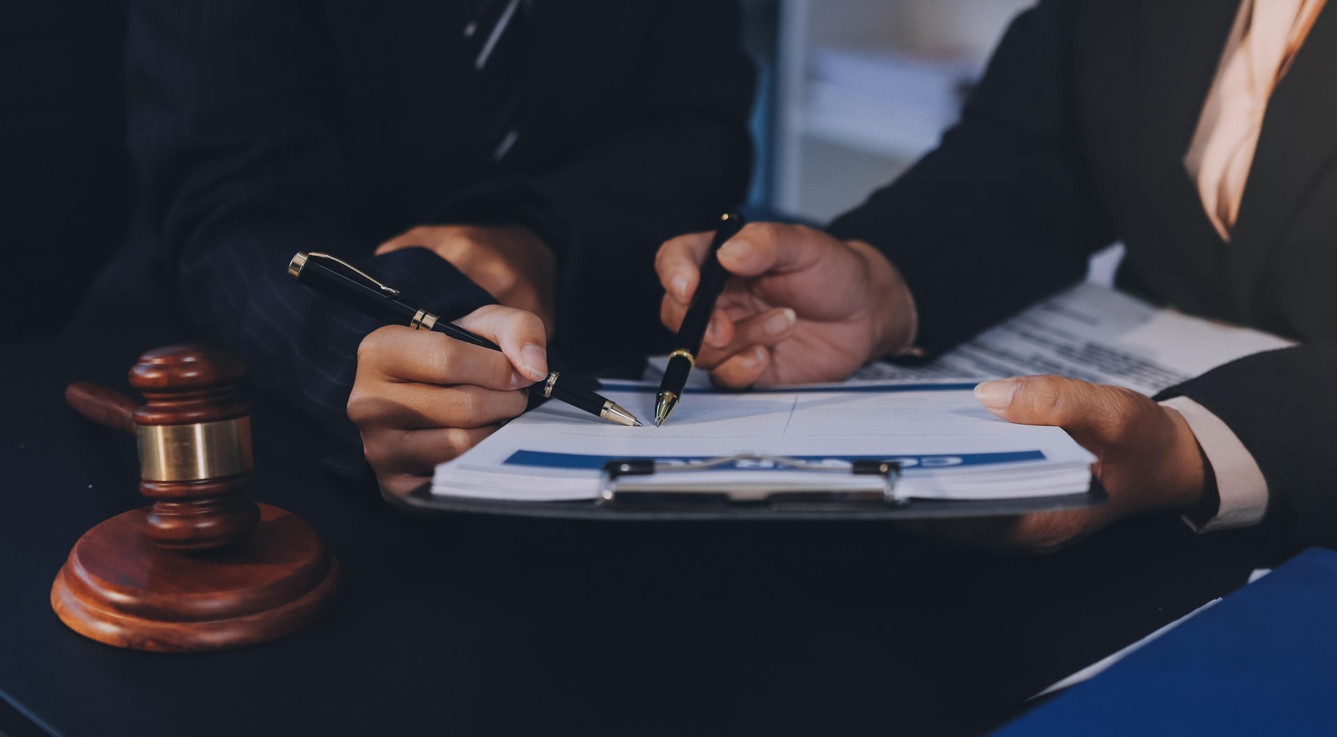 Two people in suits review documents, one signing with a pen, gavel on desk.