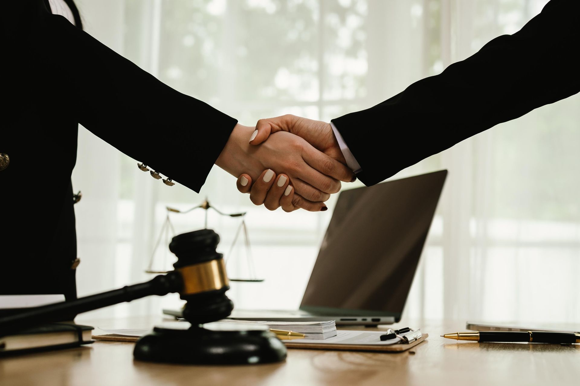 Two people in suits shaking hands at a desk with a gavel, laptop, and legal documents.
