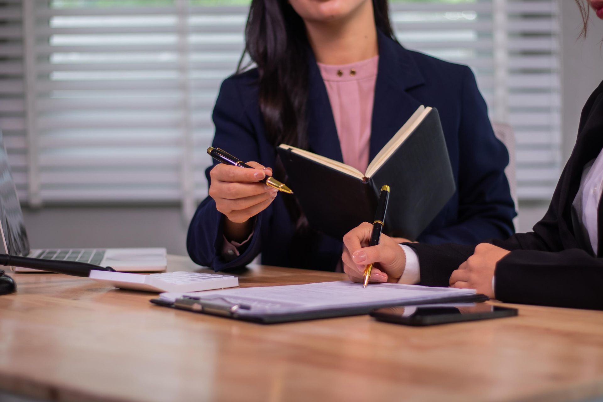 Two people in suits at a desk, one holding a pen over a document while another holds a notebook.