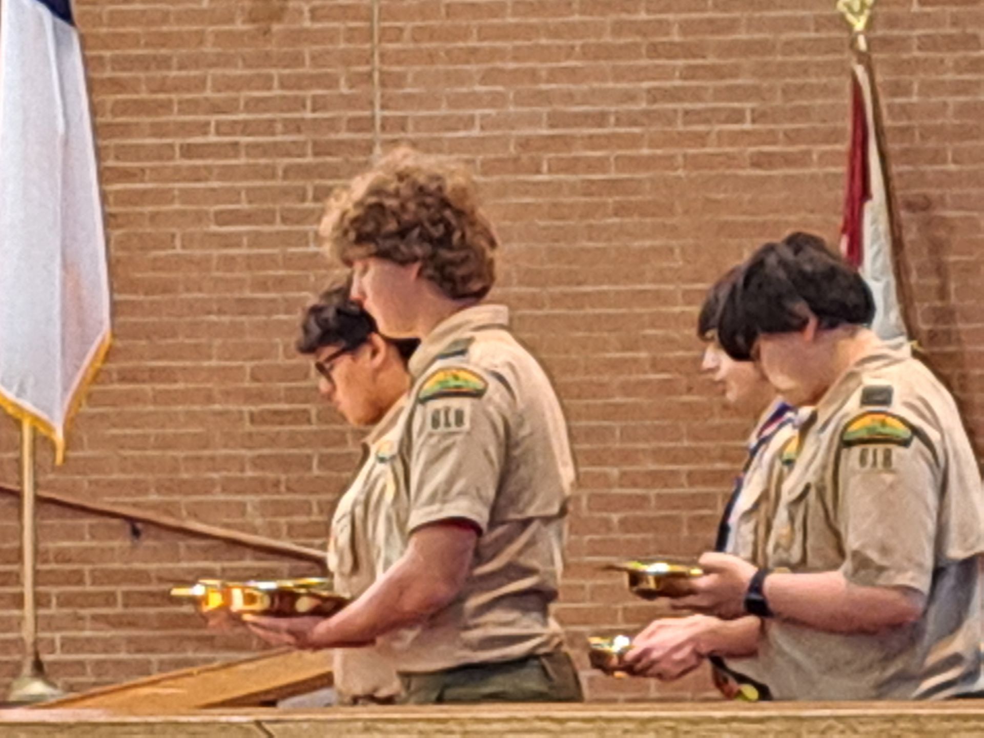 Boy Scouts holding bowls during a ceremony, in front of a brick wall and flags.