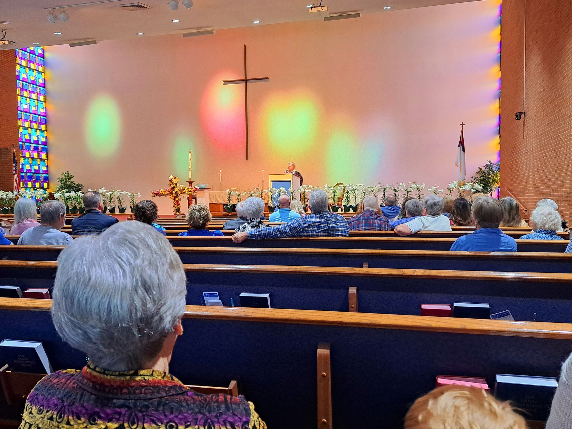 People seated in pews, facing a cross and speaker at a church service. Colorful lights illuminate the wall.