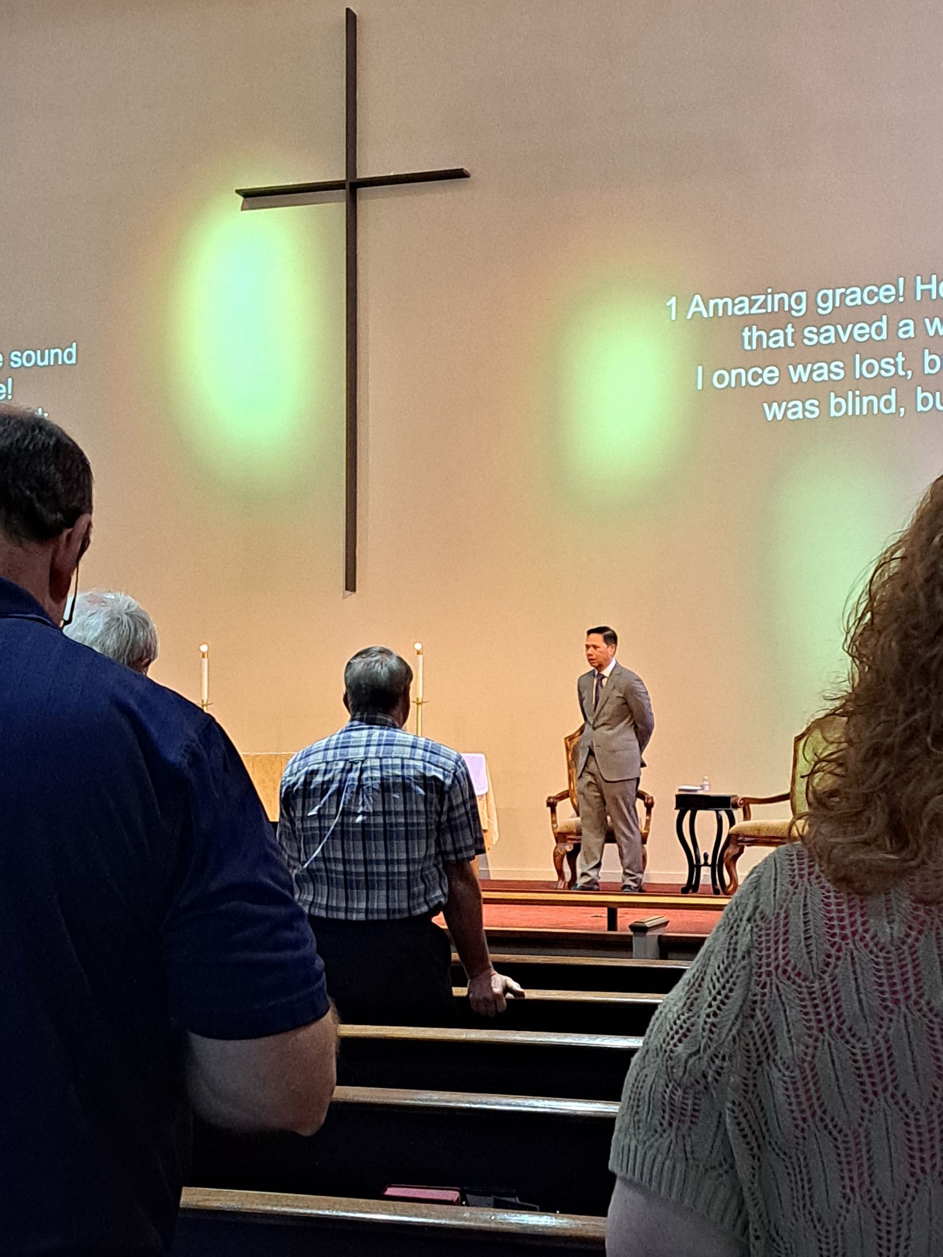People in a church service; a pastor on the stage, cross on the wall with lyrics projected.
