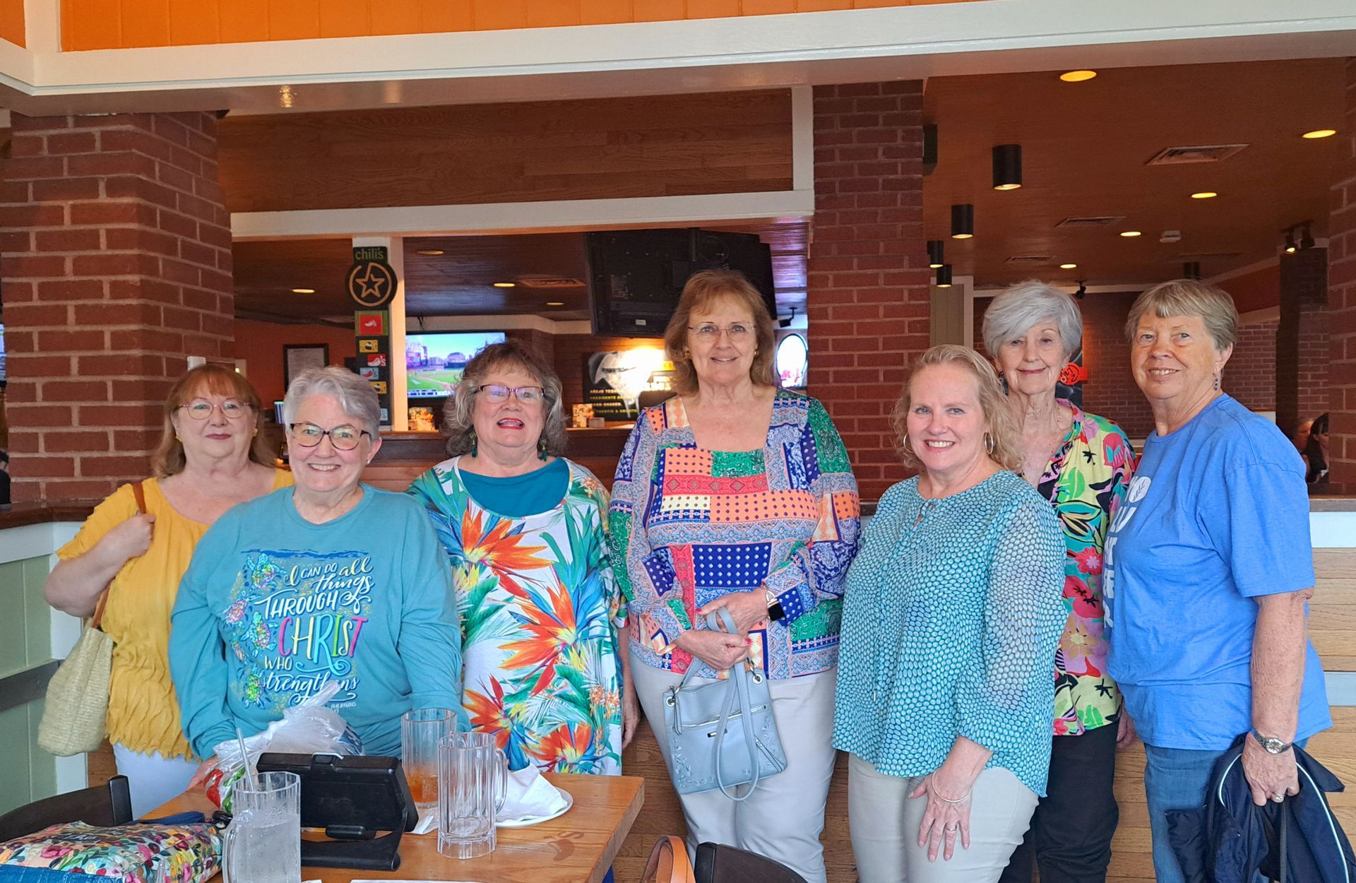 Group of women smiling, posing at a restaurant. Colorful outfits, brick wall, and overhead lighting.