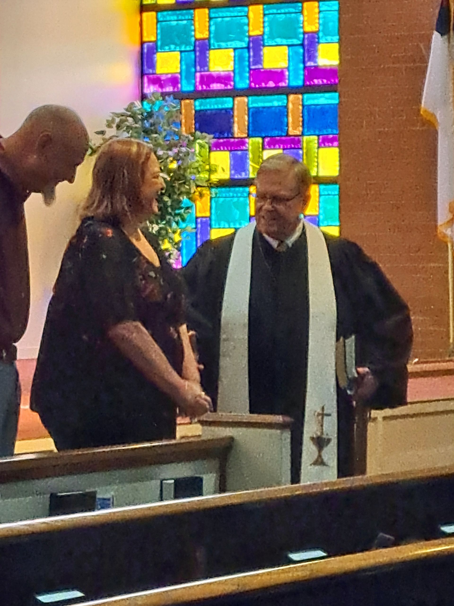 A woman in black and a man with a shaved head stand near a pastor in a church.