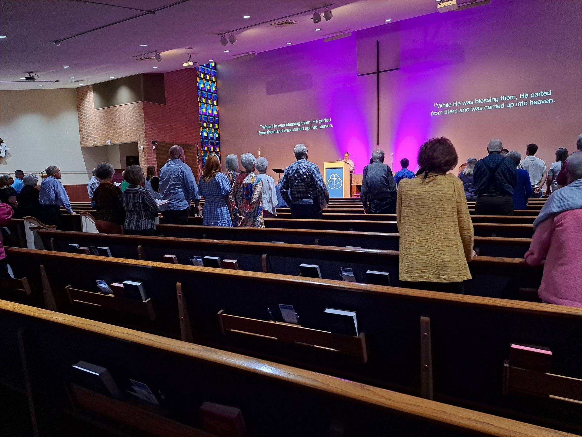 People standing in pews at a church, facing a stage with a cross, illuminated by purple light.