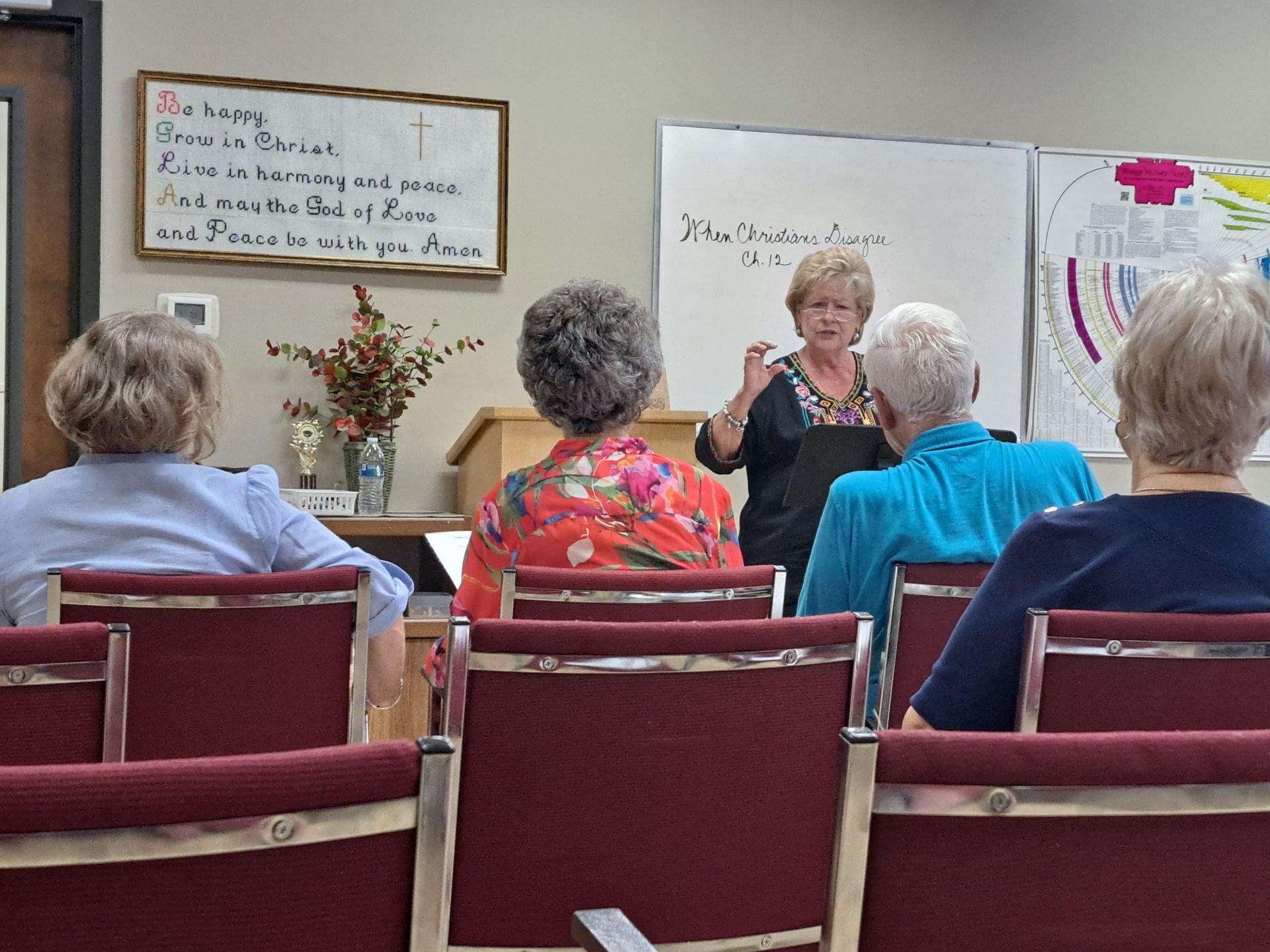 Woman lecturing to seated audience; whiteboard with writing and a diagram visible.