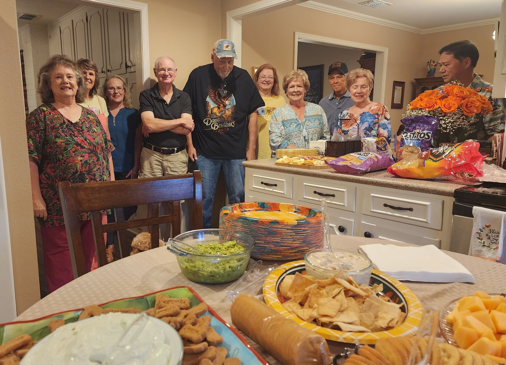 Group of people around a kitchen island with snacks; a table in the foreground has more food.