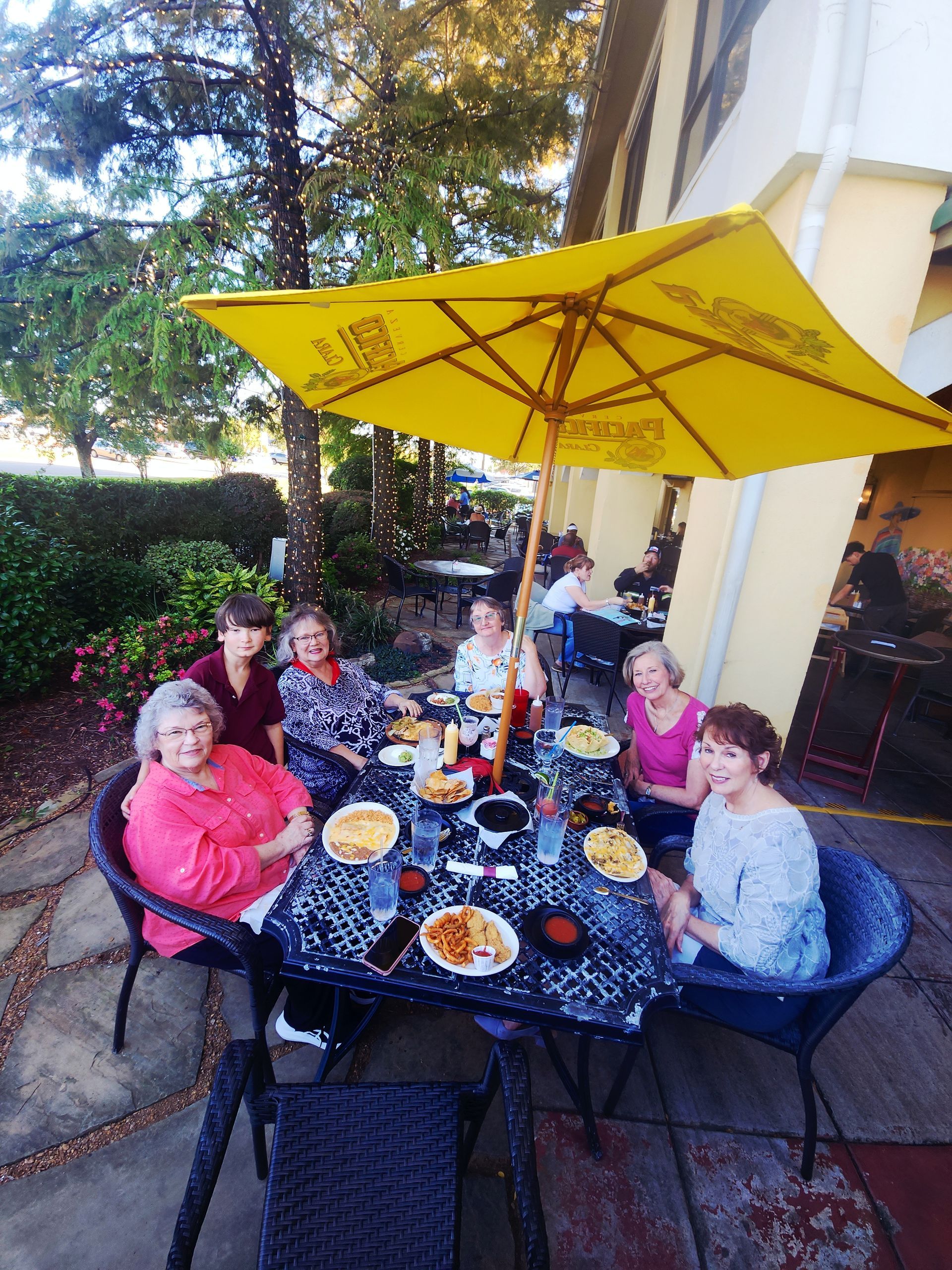 Group of people seated at an outdoor table under a yellow umbrella, enjoying a meal.