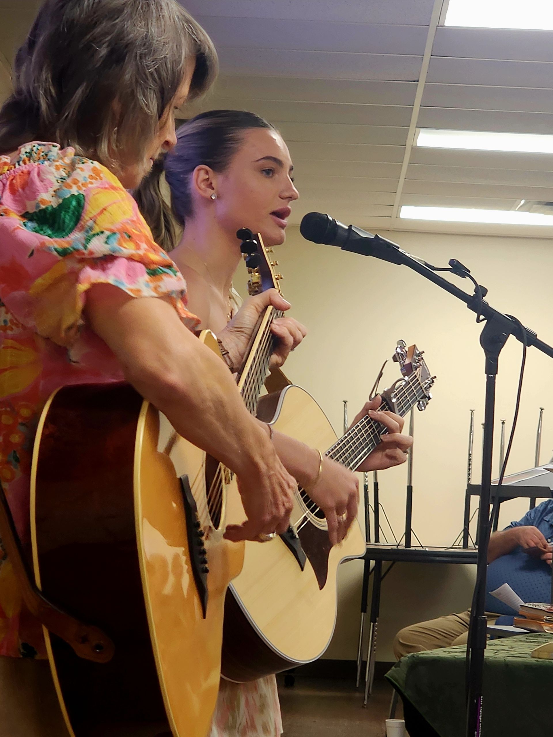 Two people playing guitars and singing into a microphone. One wears a floral top, the other a white dress.