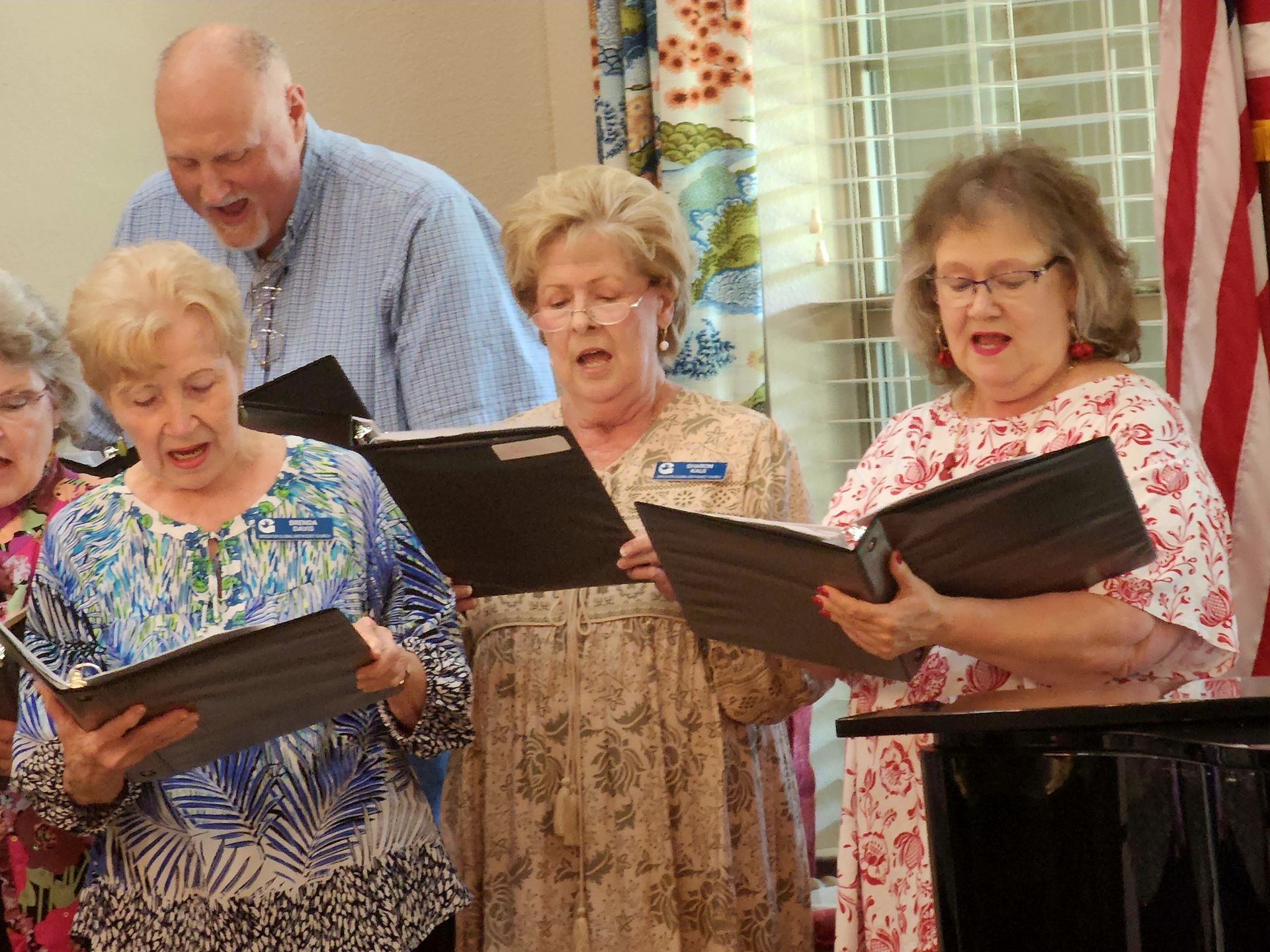Choir singing from sheet music; several people in a room, some wearing glasses and floral clothing.