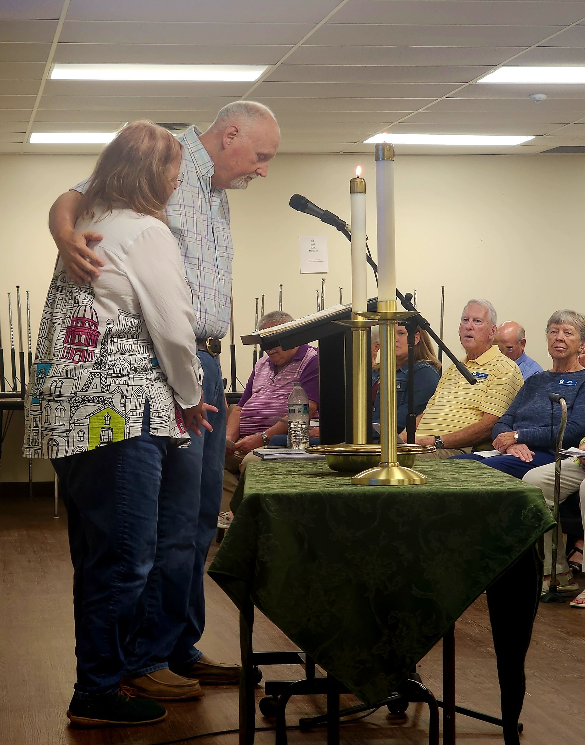 Man with arm around woman at podium, speaking to an audience. Candles and microphone present.