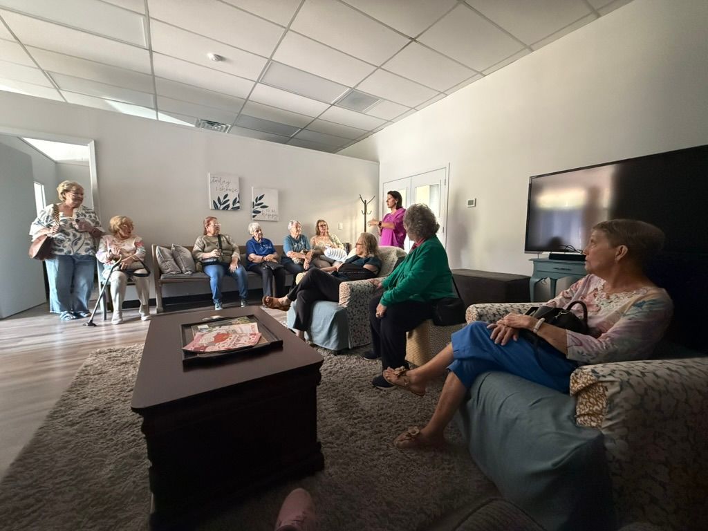 Group of people in a light-filled living room, some seated, others standing; a presentation or social gathering.