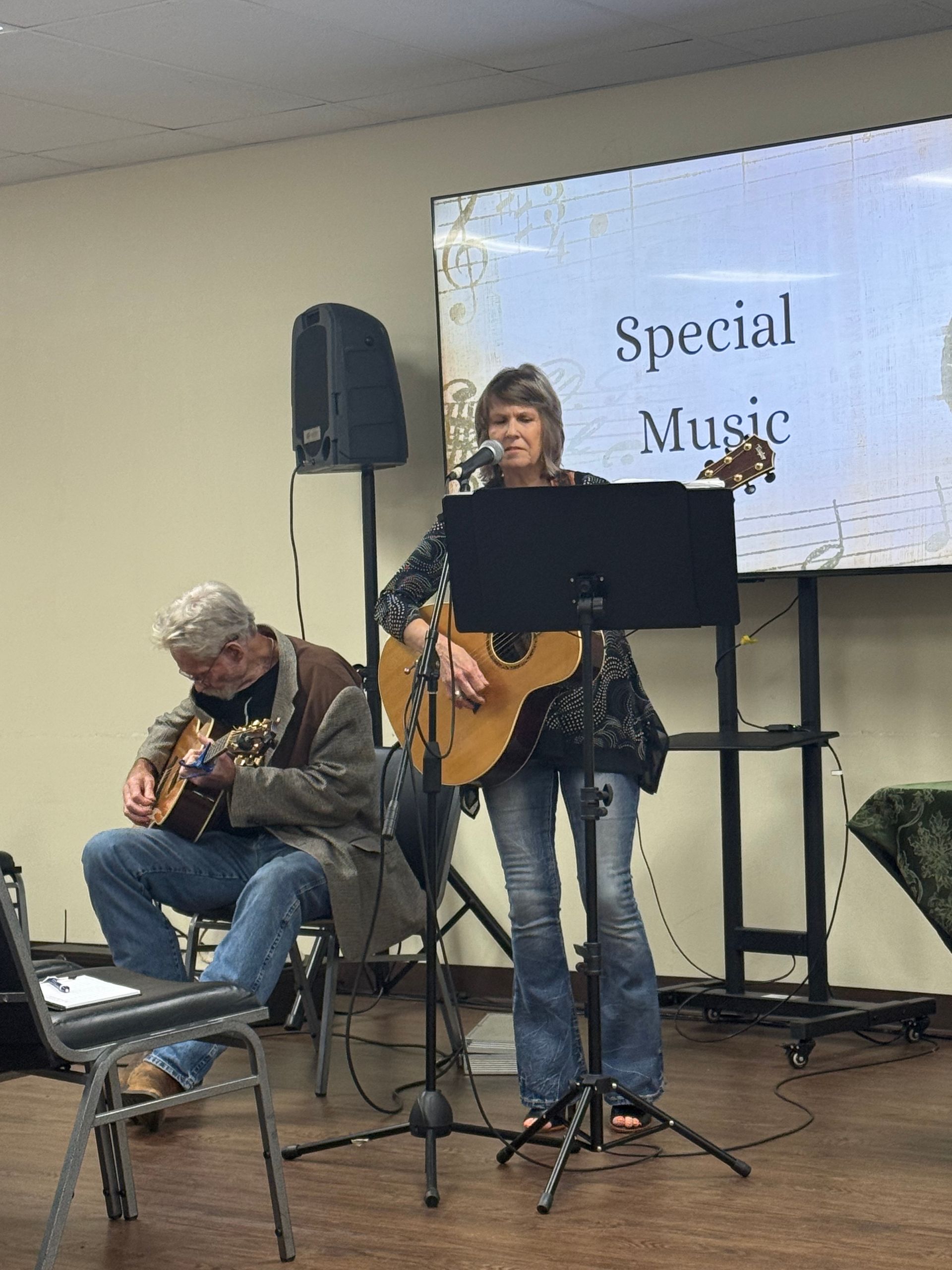 Two musicians perform at an event. A woman sings and plays guitar, while a man plays a guitar seated.