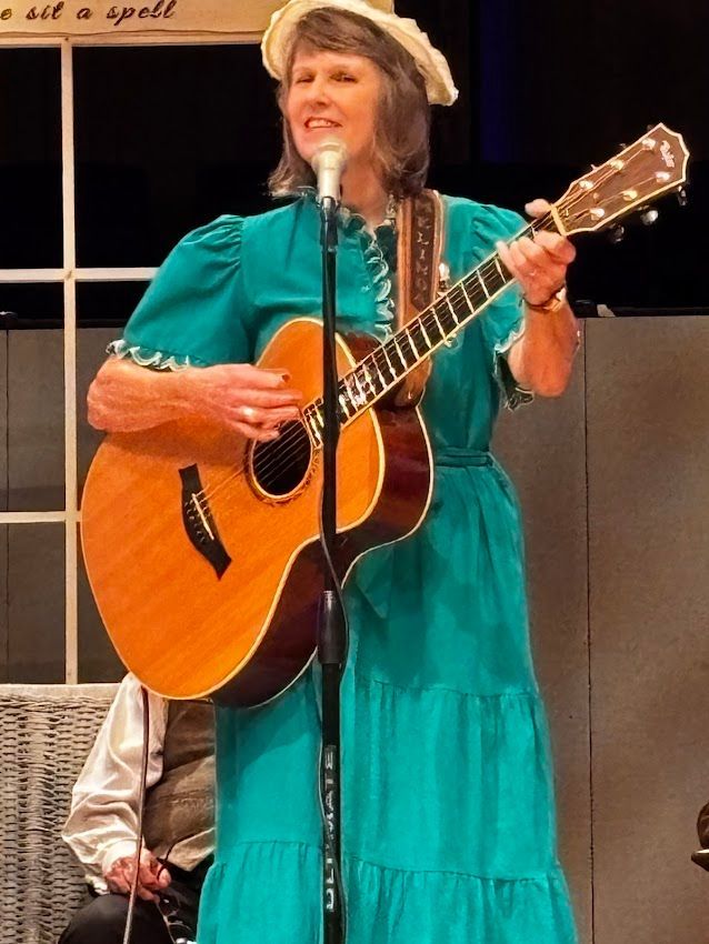 Woman in teal dress, hat, playing acoustic guitar, singing into a microphone on a stage.