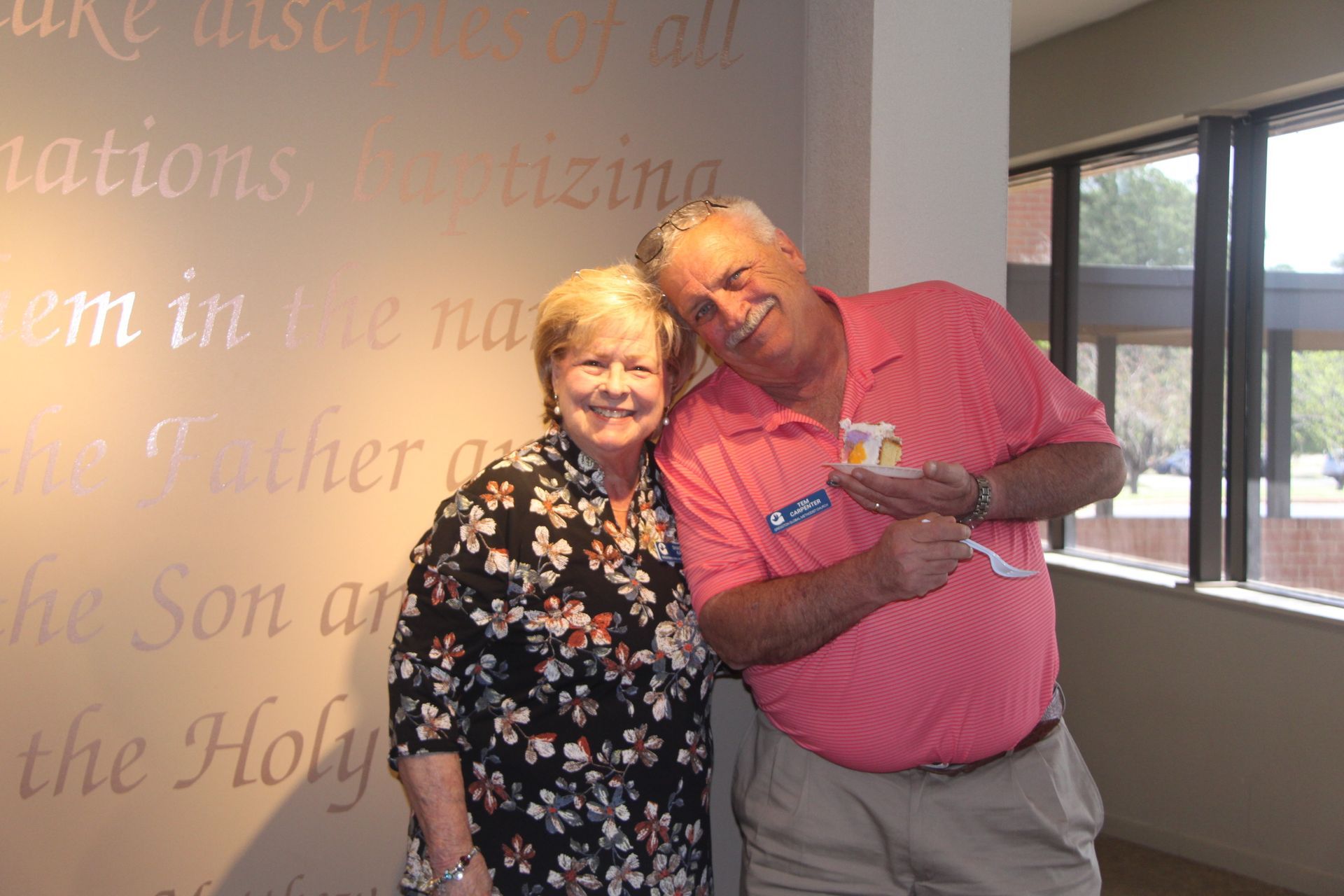 Woman and man smile, holding a cake. They stand by a wall with script. Man wears pink shirt, woman floral blouse.