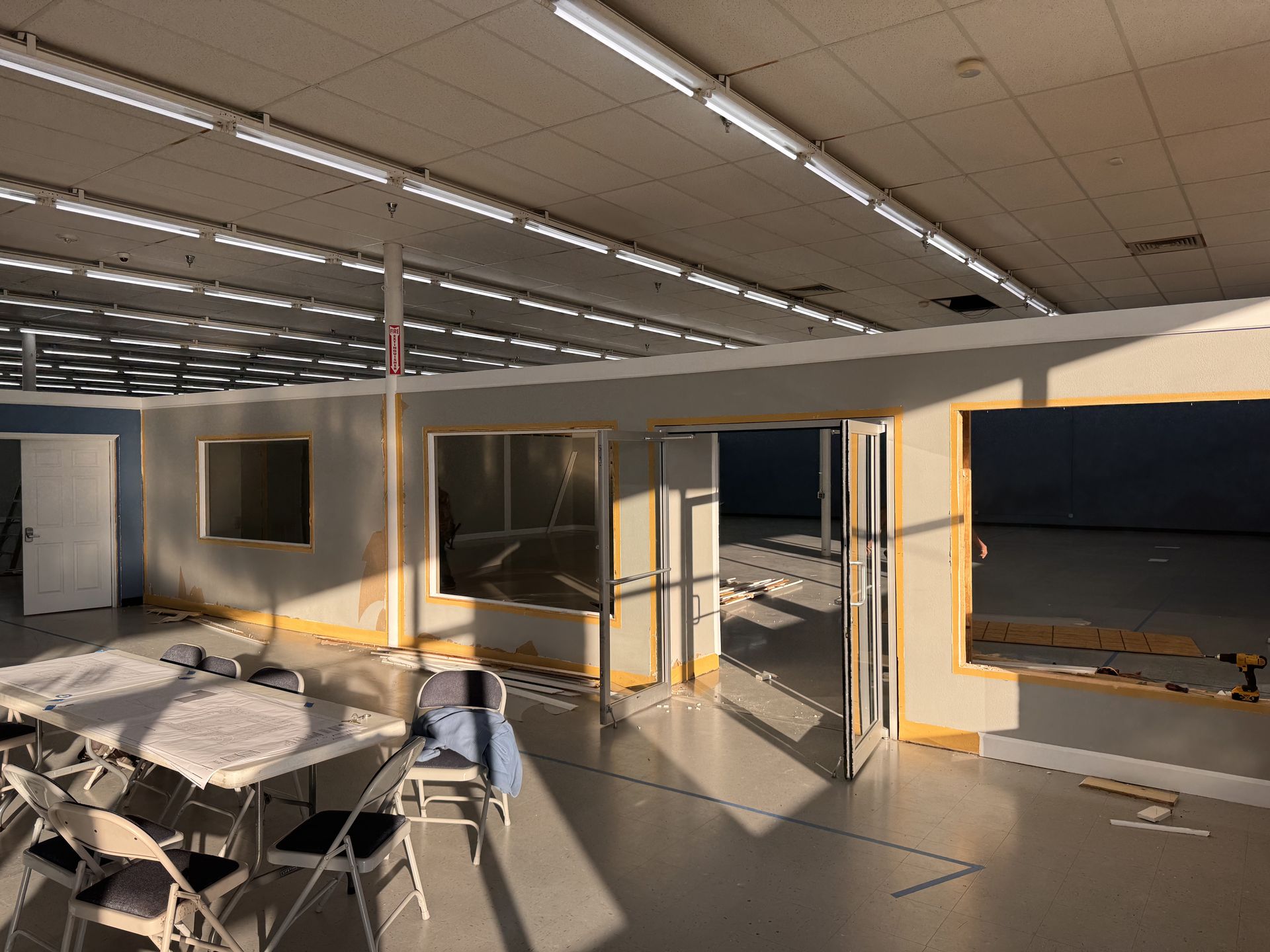 Interior construction: Walls with windows and doorway in a large, open space. Fluorescent lighting. Tables and chairs in foreground.