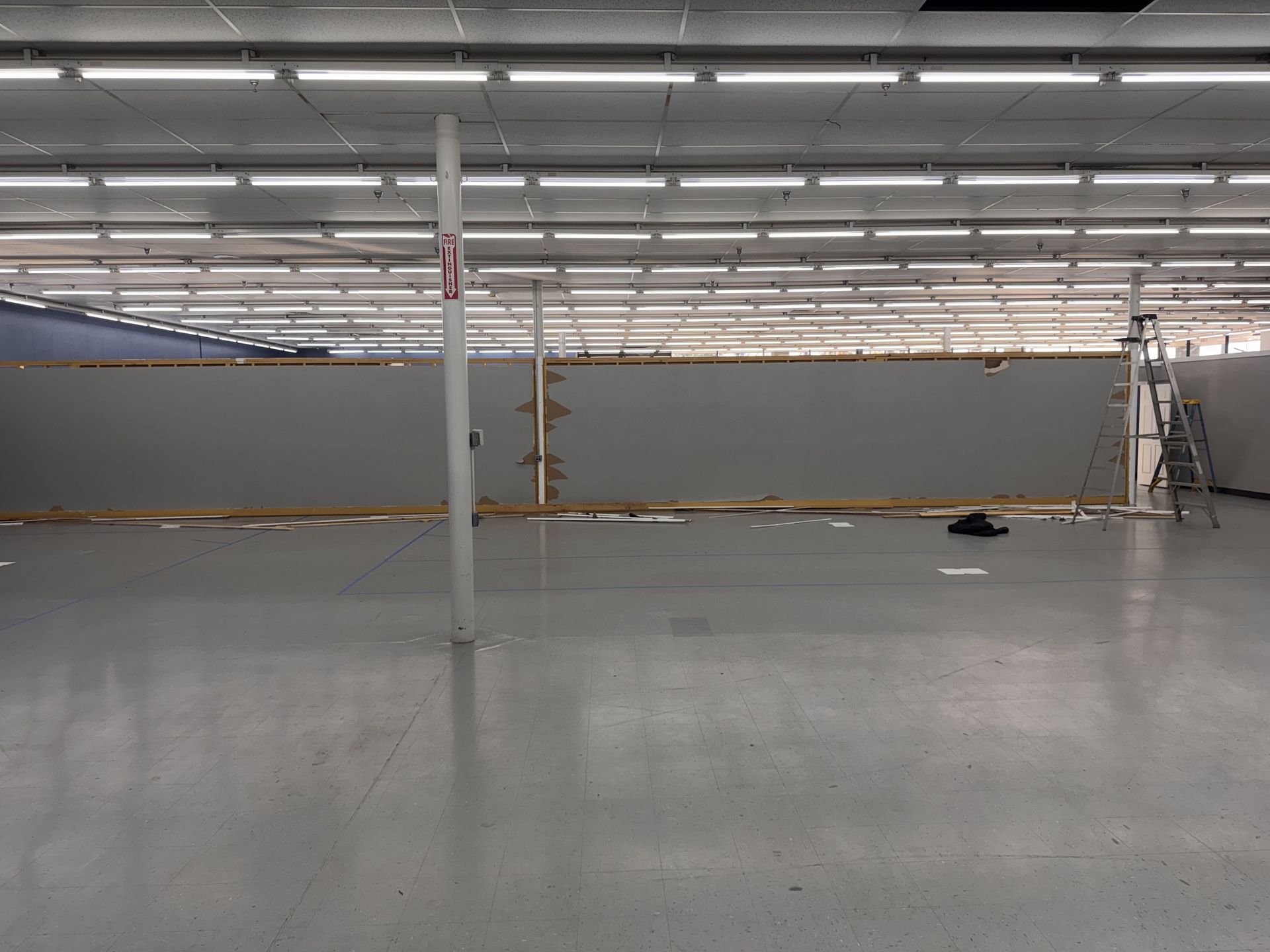 Empty store interior with gray floors, white ceiling, and gray wall in the background. A pole and ladder are visible.