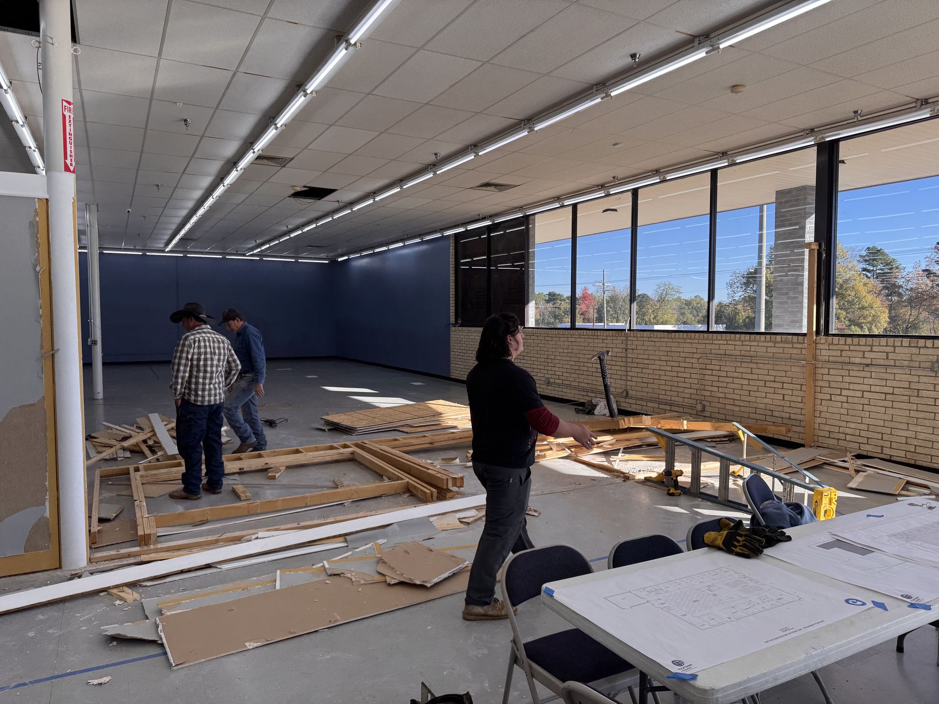 Interior construction site: Workers dismantling walls and framework. Debris litters floor near windows and tables.