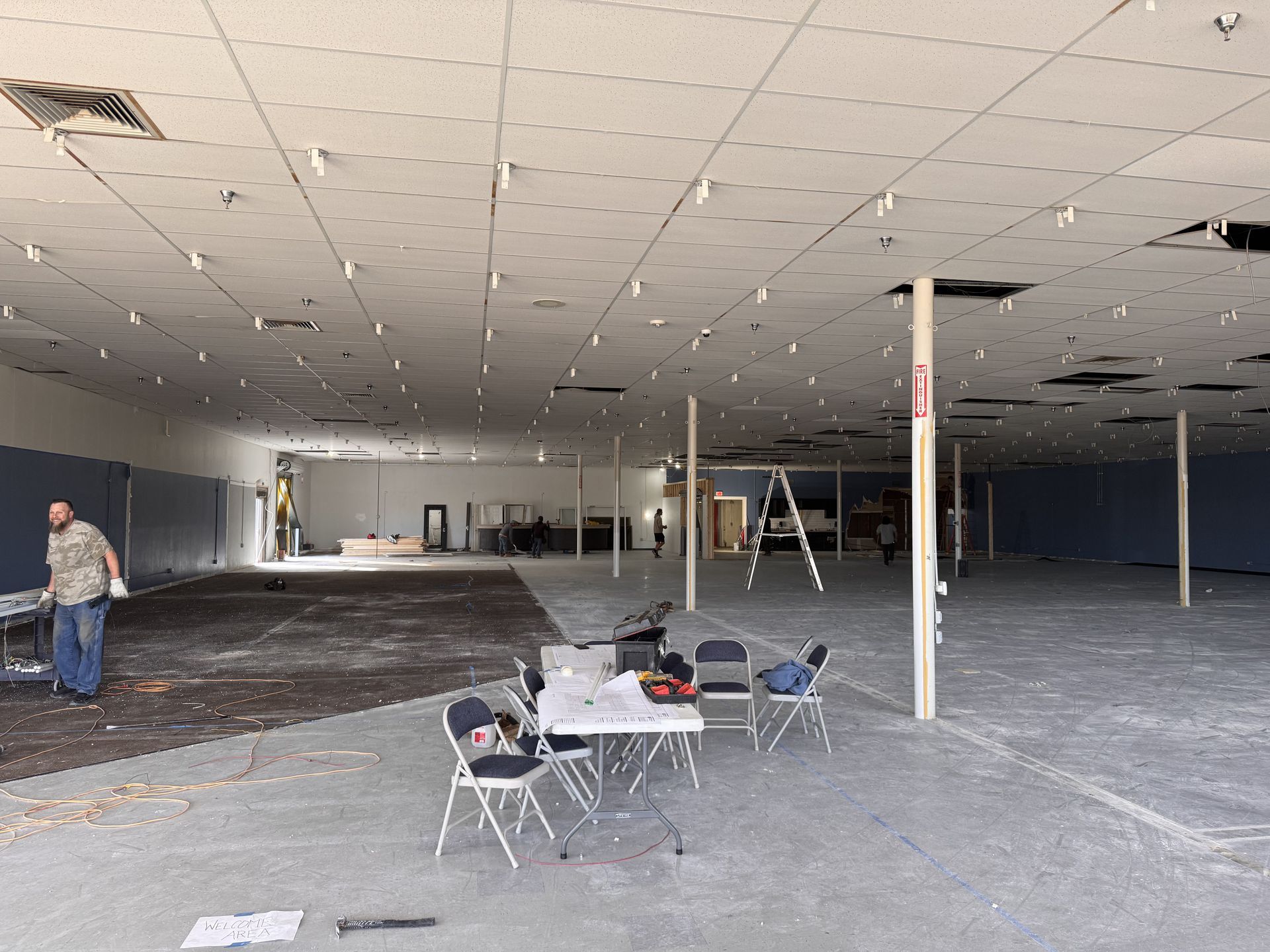 Empty commercial space under renovation, with workers, tables, and supplies. Gray and white interior.