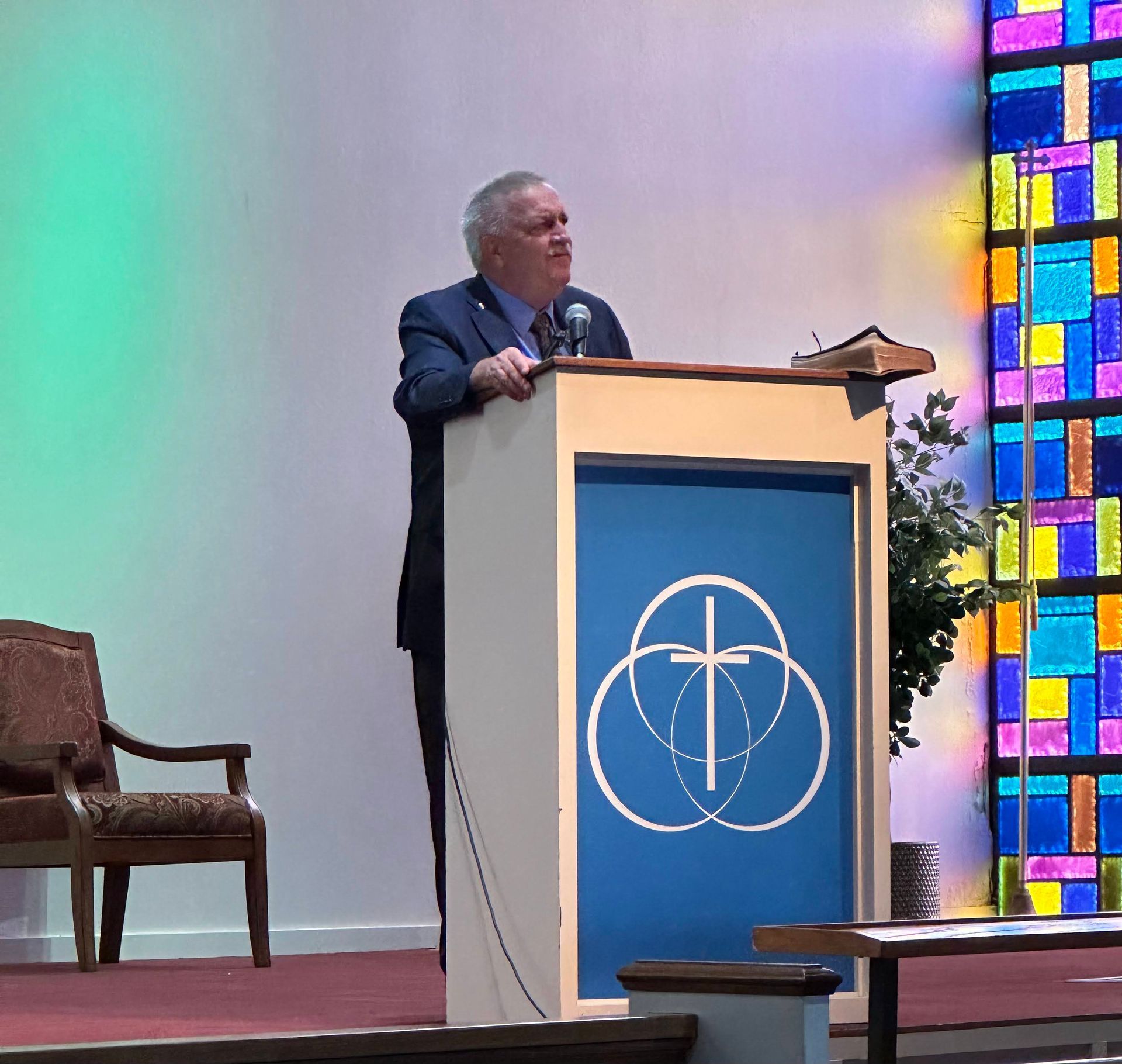 Man speaking from a podium with a church logo, near stained glass.