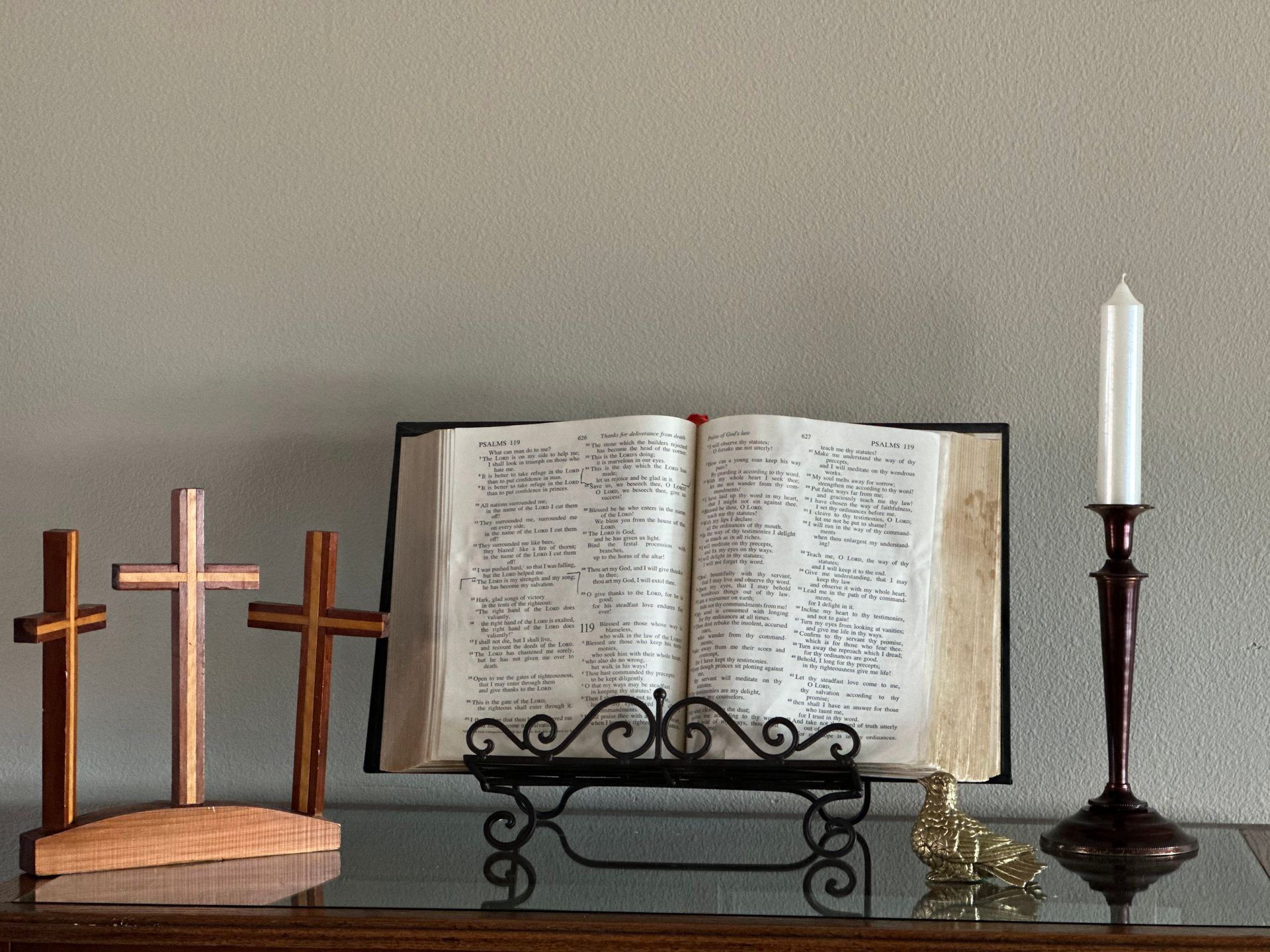 Three wooden crosses, open book on stand, candle in holder. On a dark surface against a gray wall.